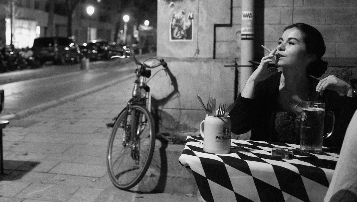 A woman sits at a cafe at night with a beer and a cigarette