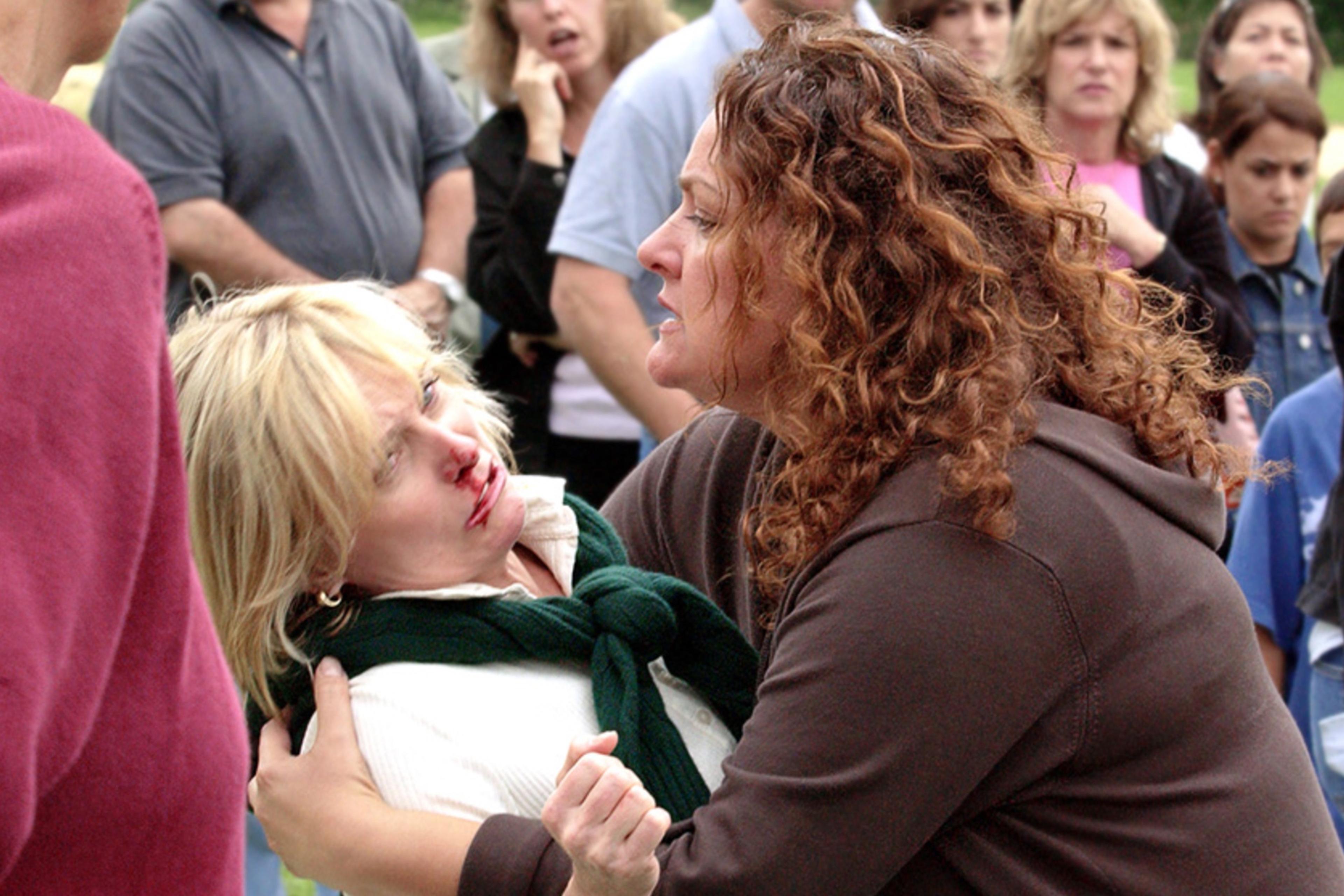 Photo of a woman with curly hair comforting an injured woman in a crowd, whose nose is bleeding.
