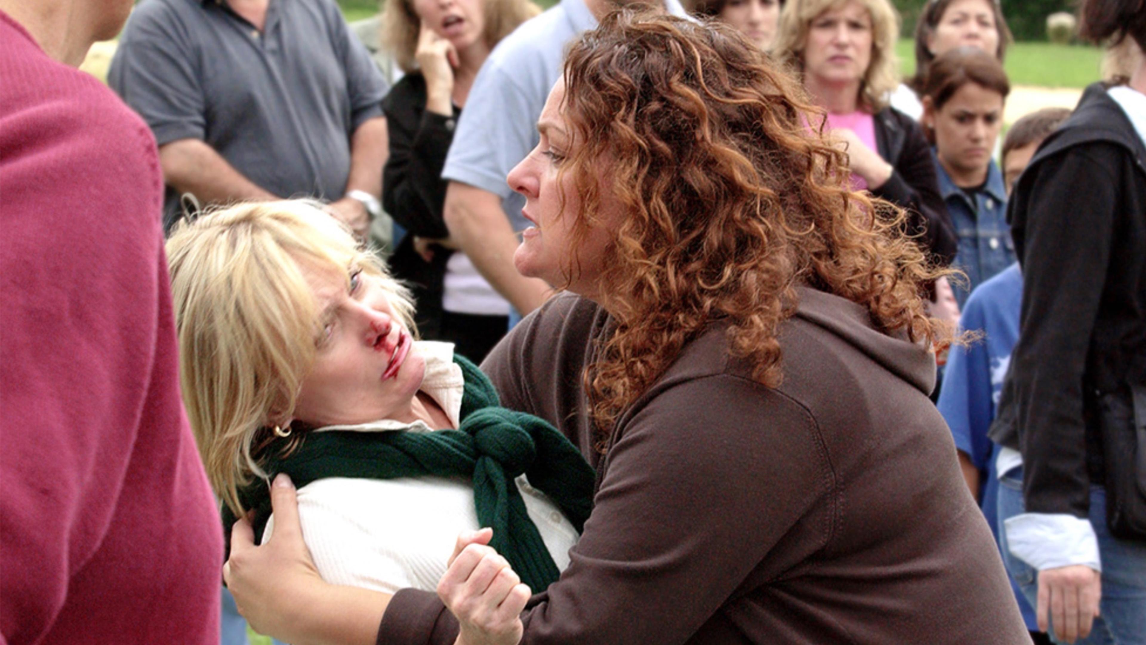 Photo of a woman with curly hair comforting an injured woman in a crowd, whose nose is bleeding.
