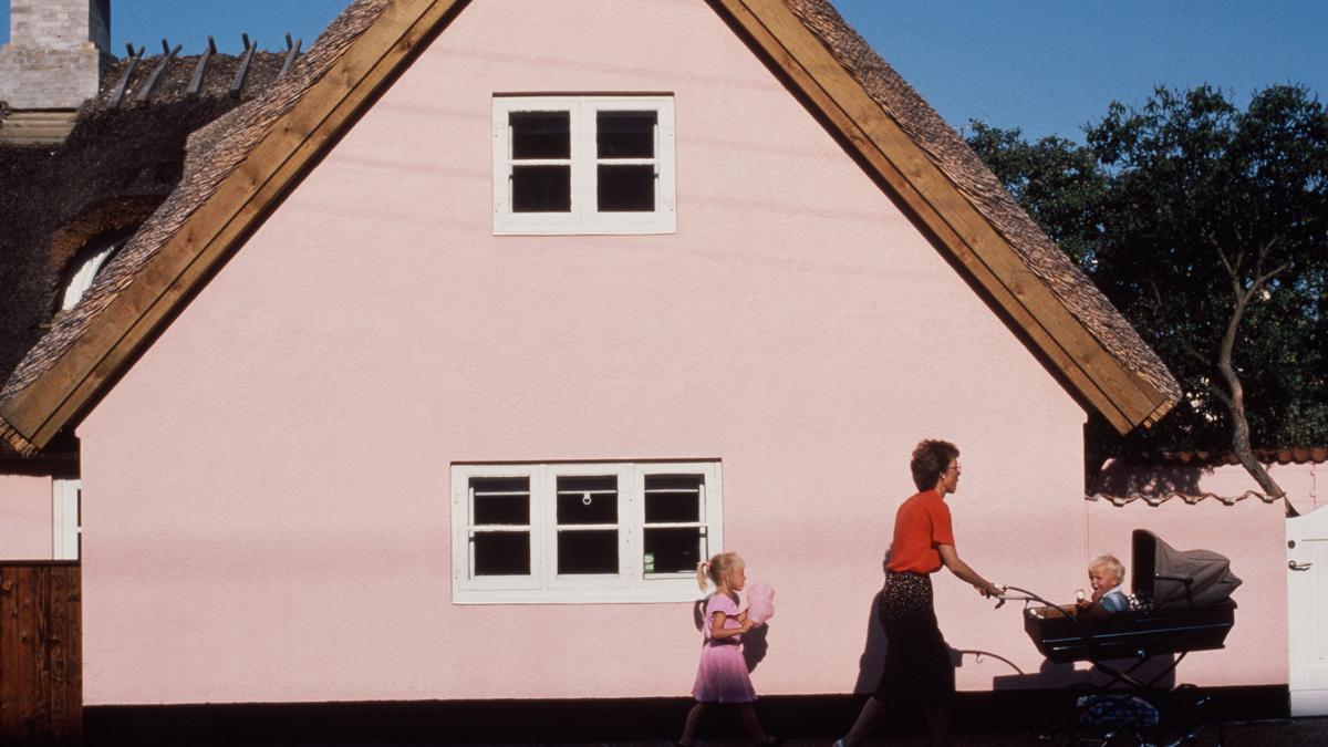 A pink cottage with a thatched roof; a woman pushes a baby in a pram alongside a girl holding candyfloss.