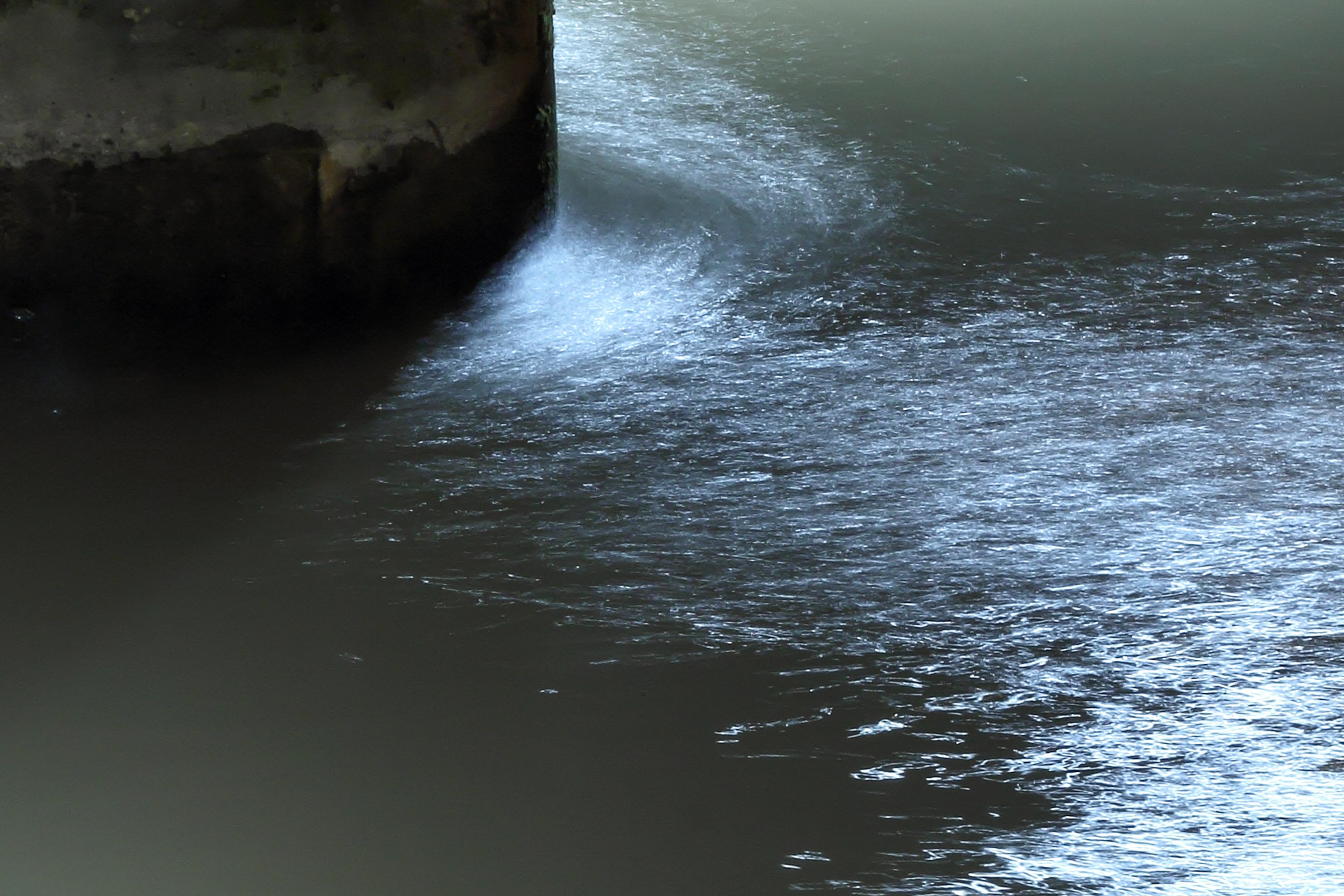 Water swirling around a concrete structure in low light, creating a reflective and slightly turbulent surface.