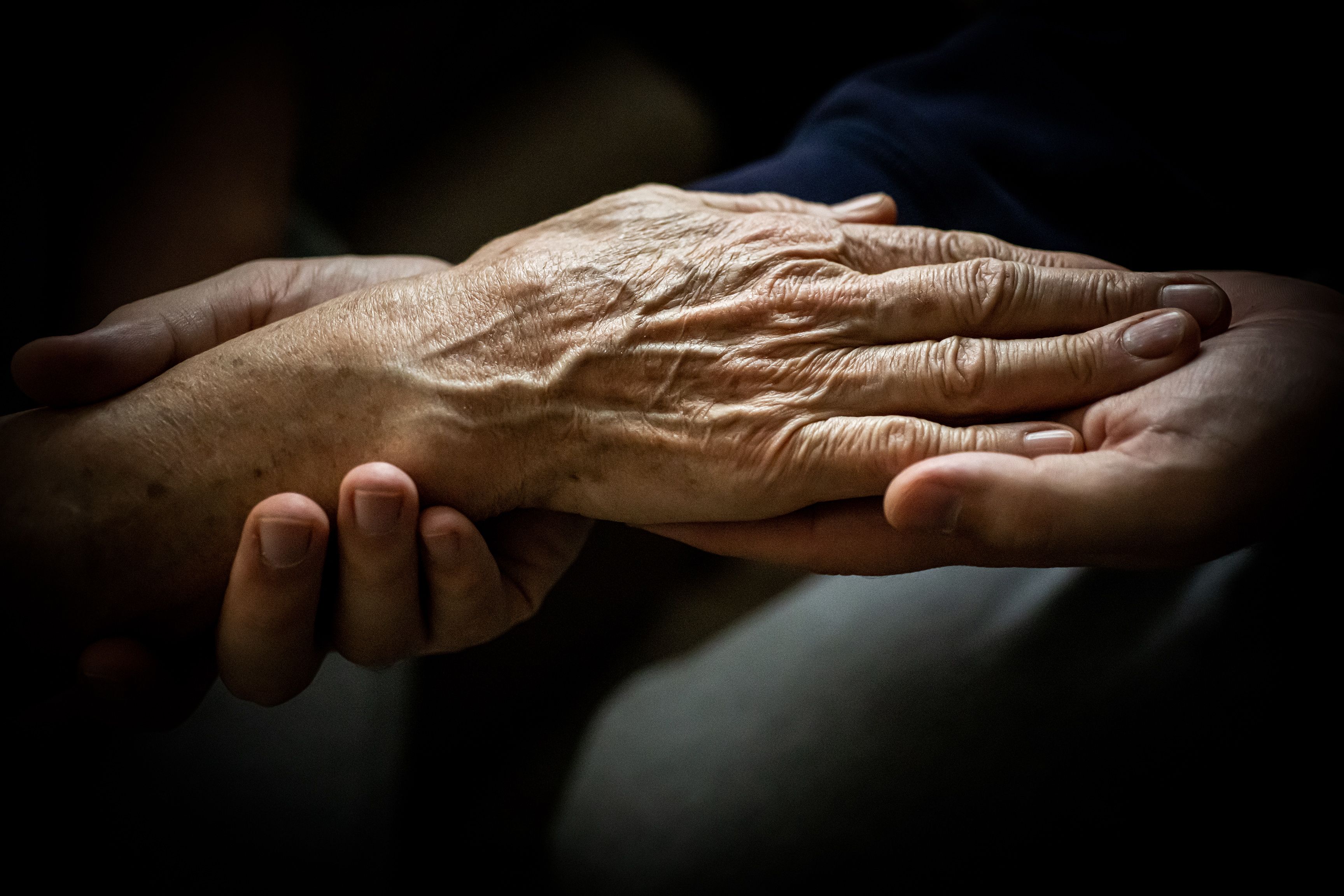 An elderly hand resting being held by younger hands against a dark background.