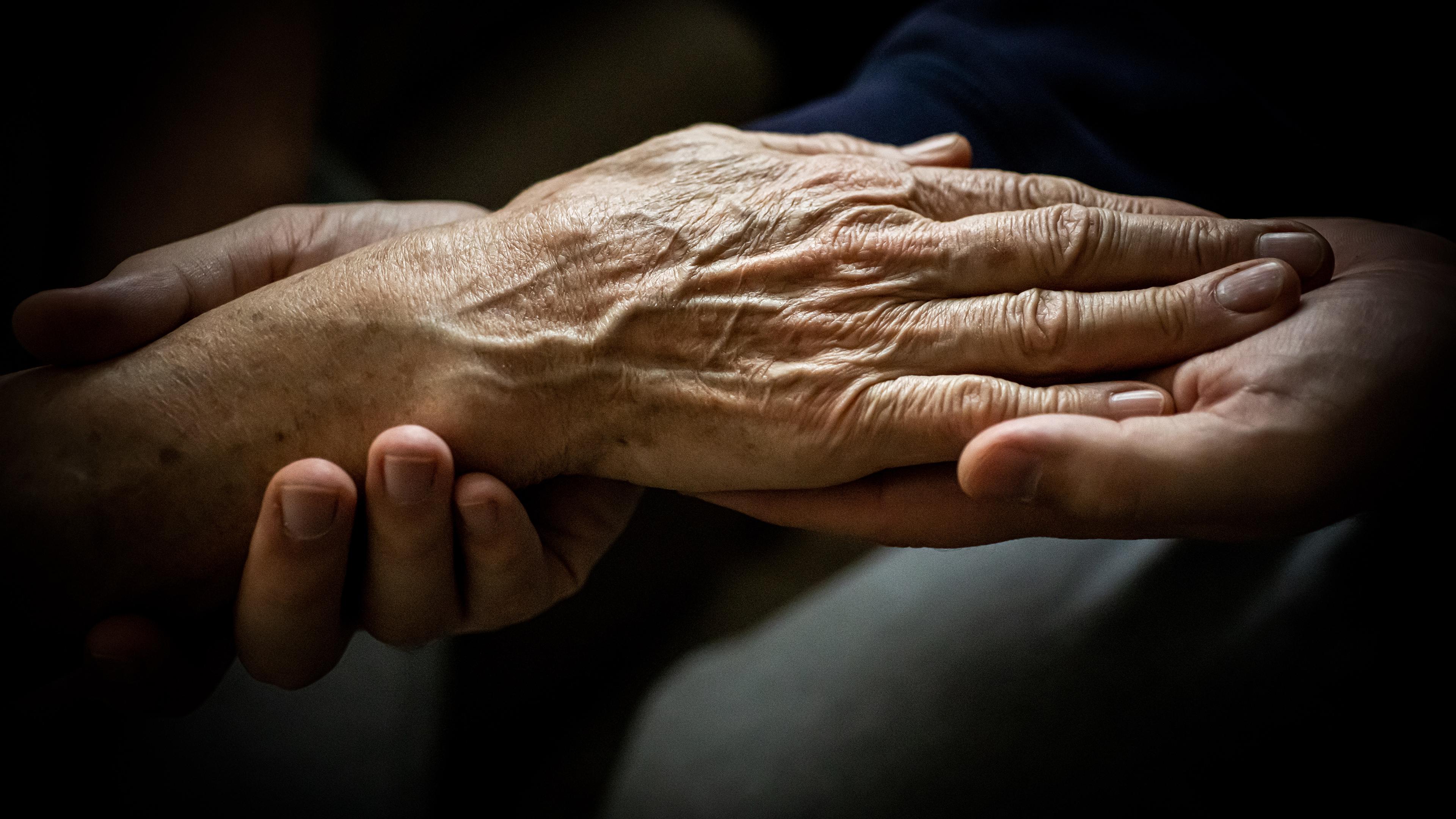An elderly hand resting being held by younger hands against a dark background.