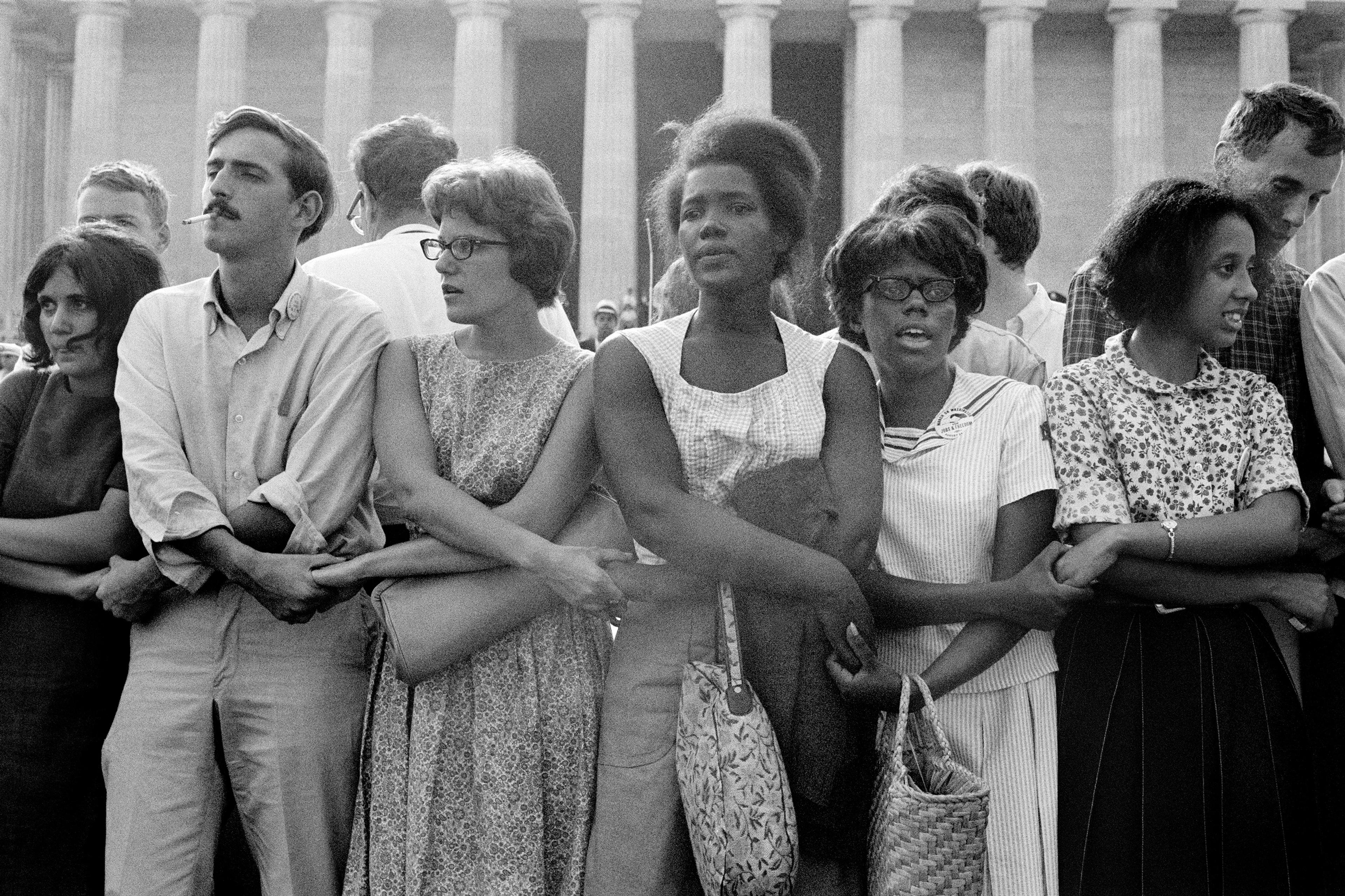 Black and white photo of a diverse group of people standing arm in arm in front of a columned building, showing solidarity.