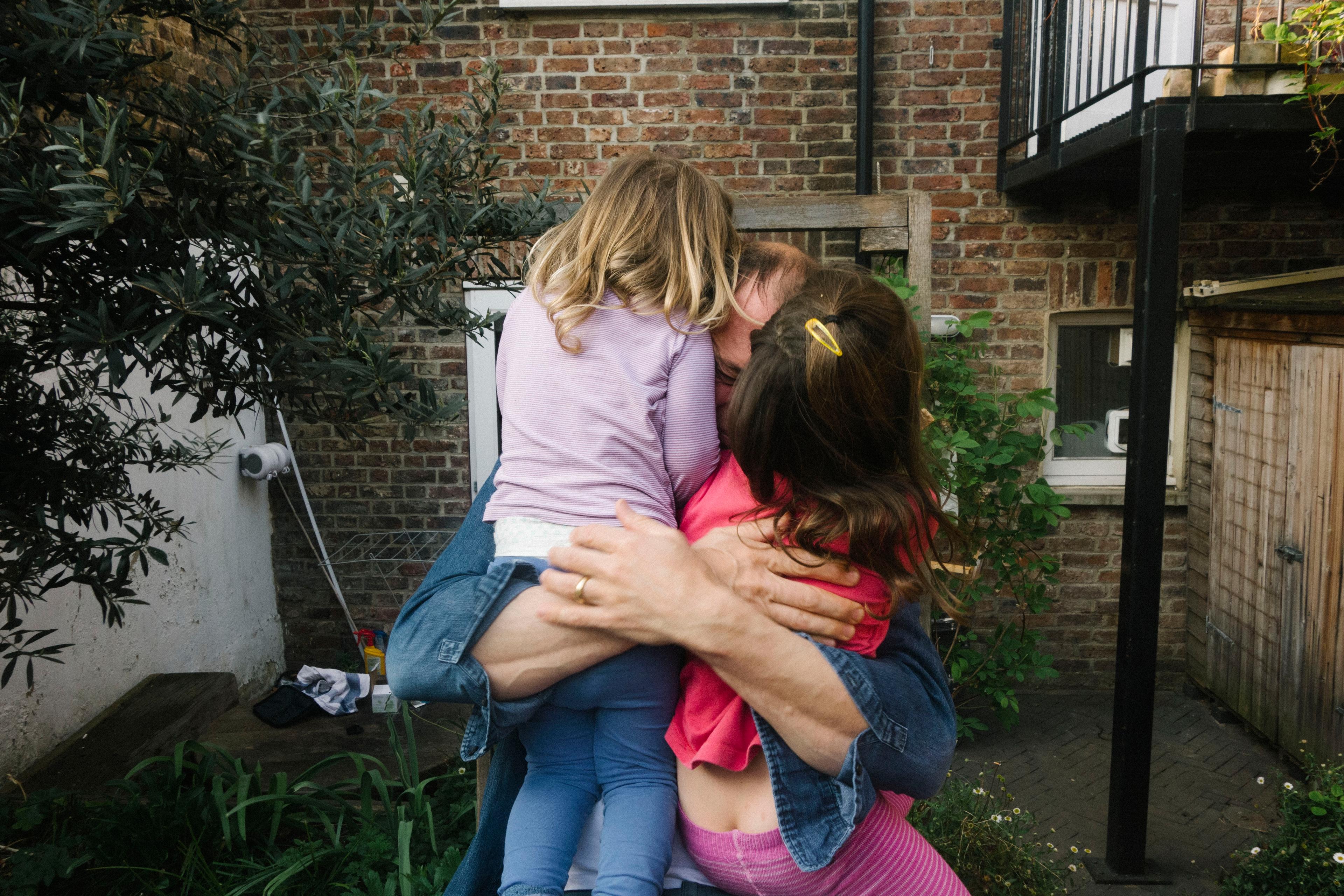 Photo of a person hugging two children in an outdoor area with a brick wall background and foliage on the left.