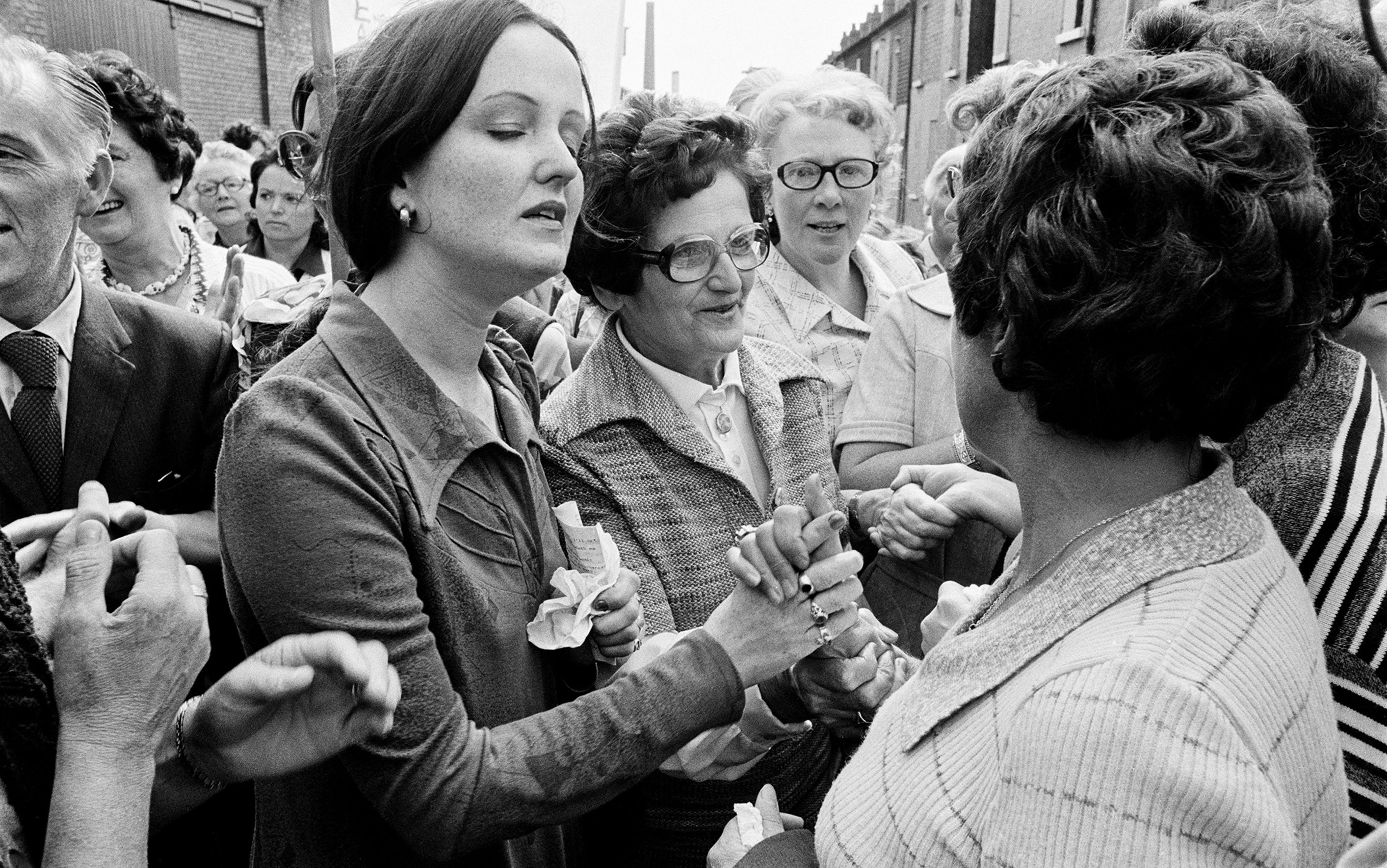 Black-and-white photo of a large gathering a street, with several women holding hands and engaging in conversation.