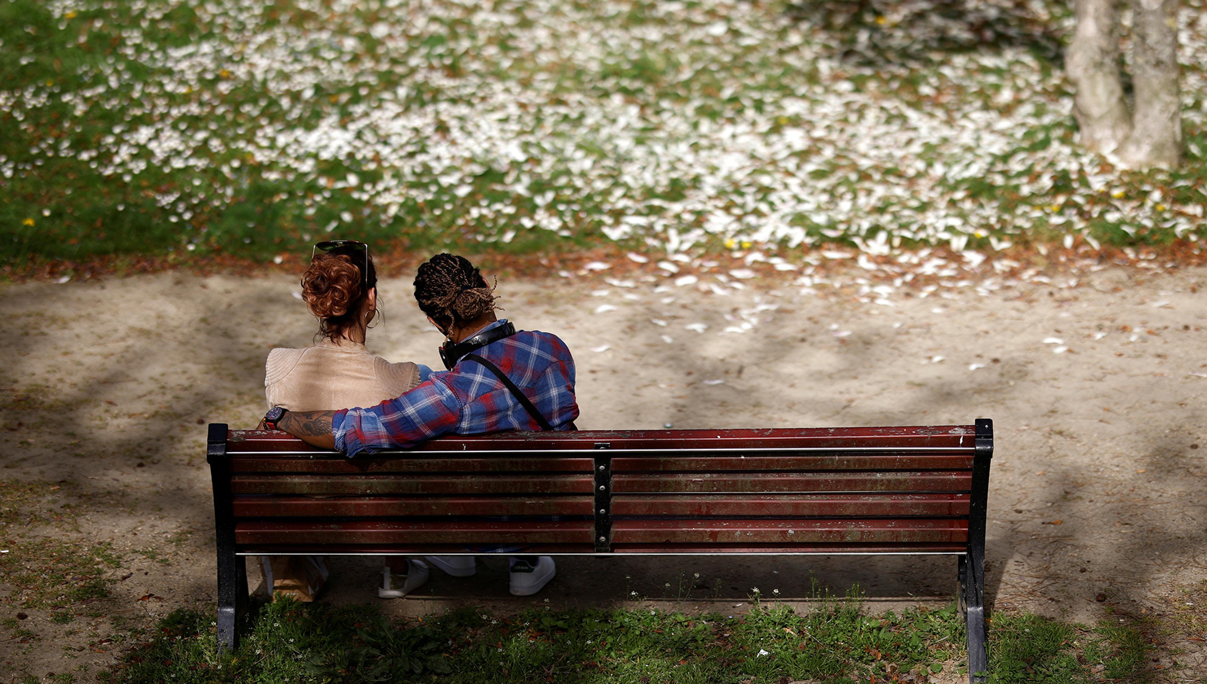 Two people sit on a wooden park bench, one with an arm around the other, with fallen leaves scattered on the ground in front of them.