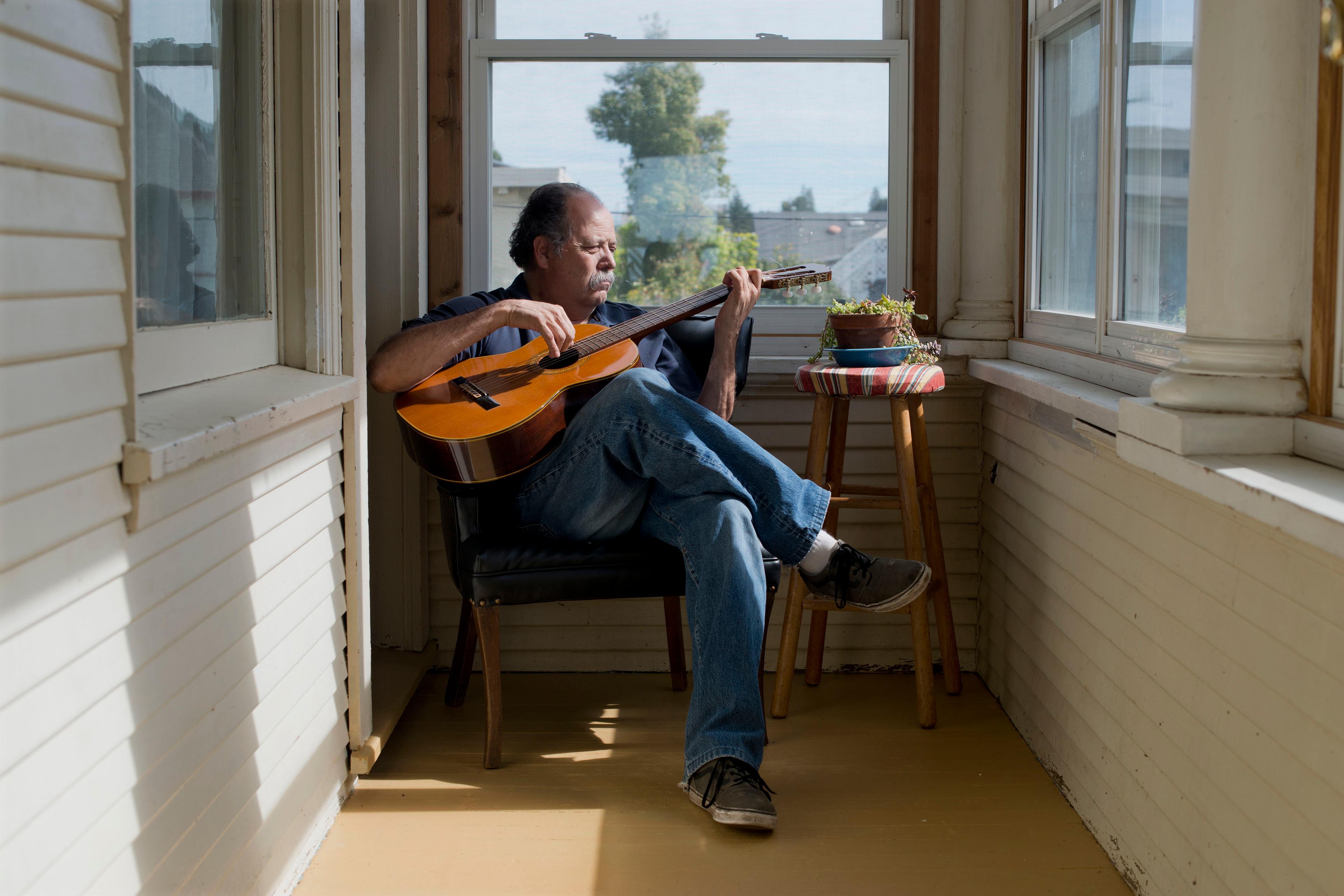 Photo of a man playing guitar in a sunlit room with wooden walls a stool with plants nearby and a view outside the window.