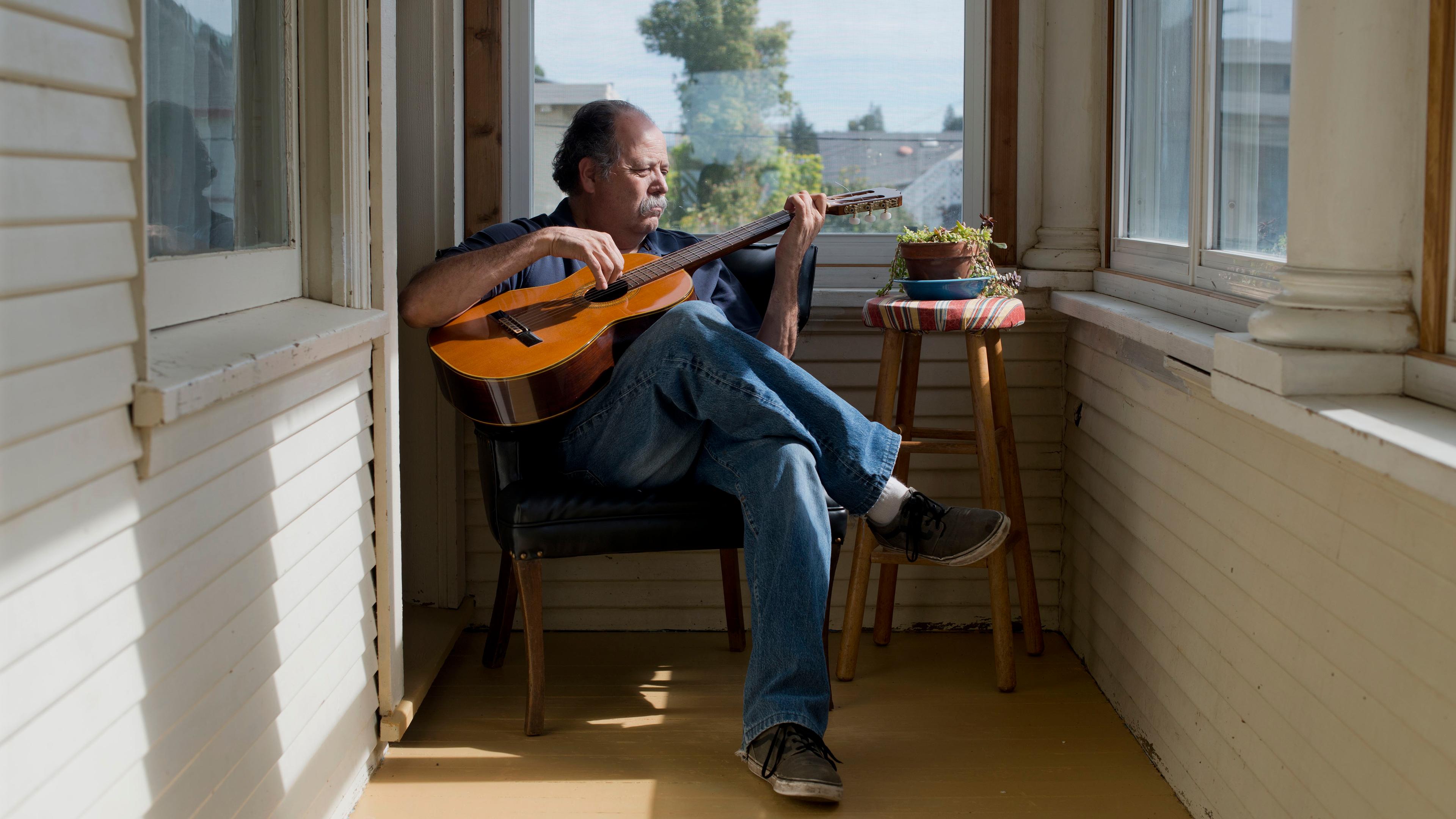 Photo of a man playing guitar in a sunlit room with wooden walls a stool with plants nearby and a view outside the window.
