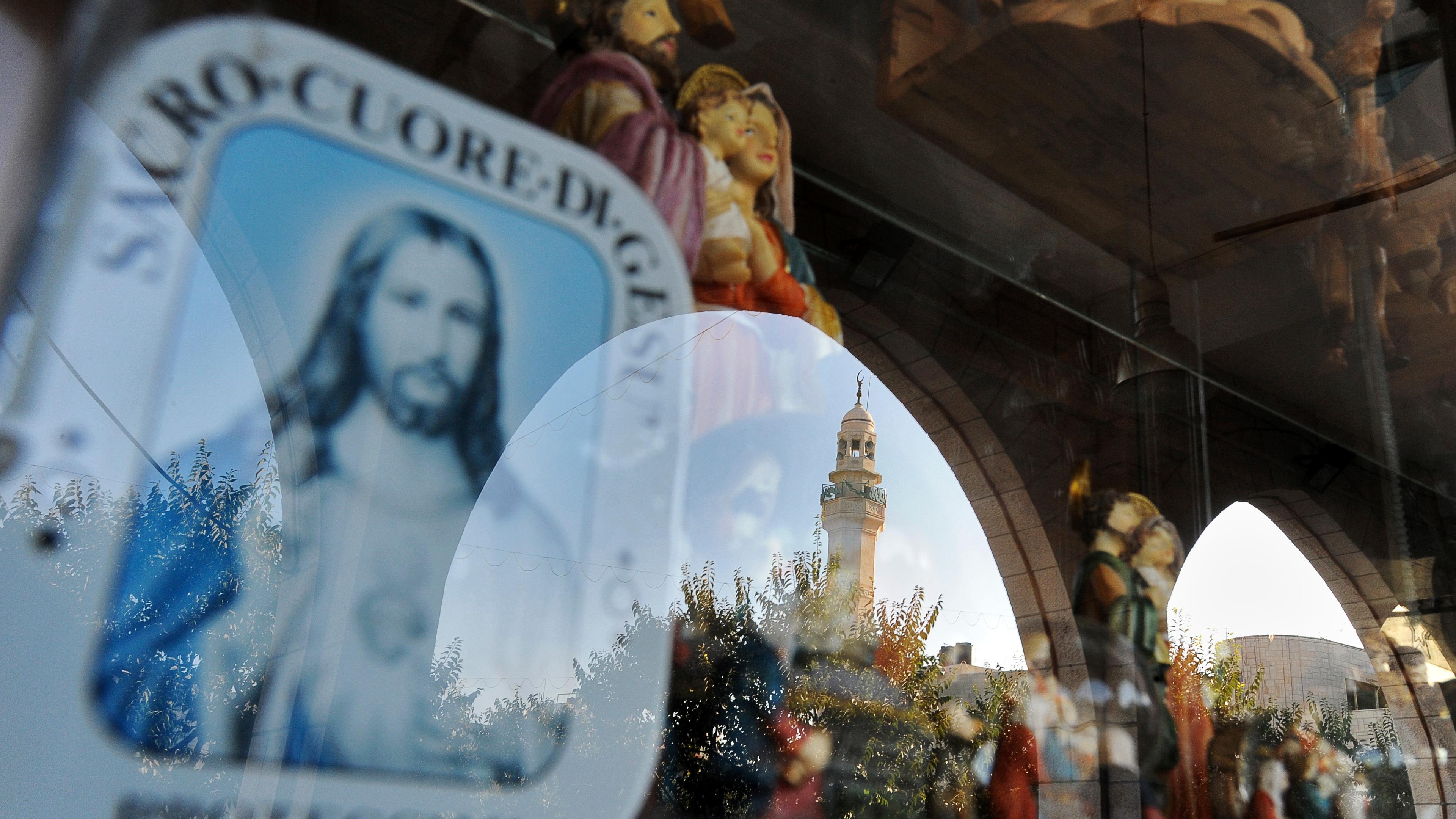 Photo of religious statues in a shop window with a reflection of an arch and a mosque tower in the background.
