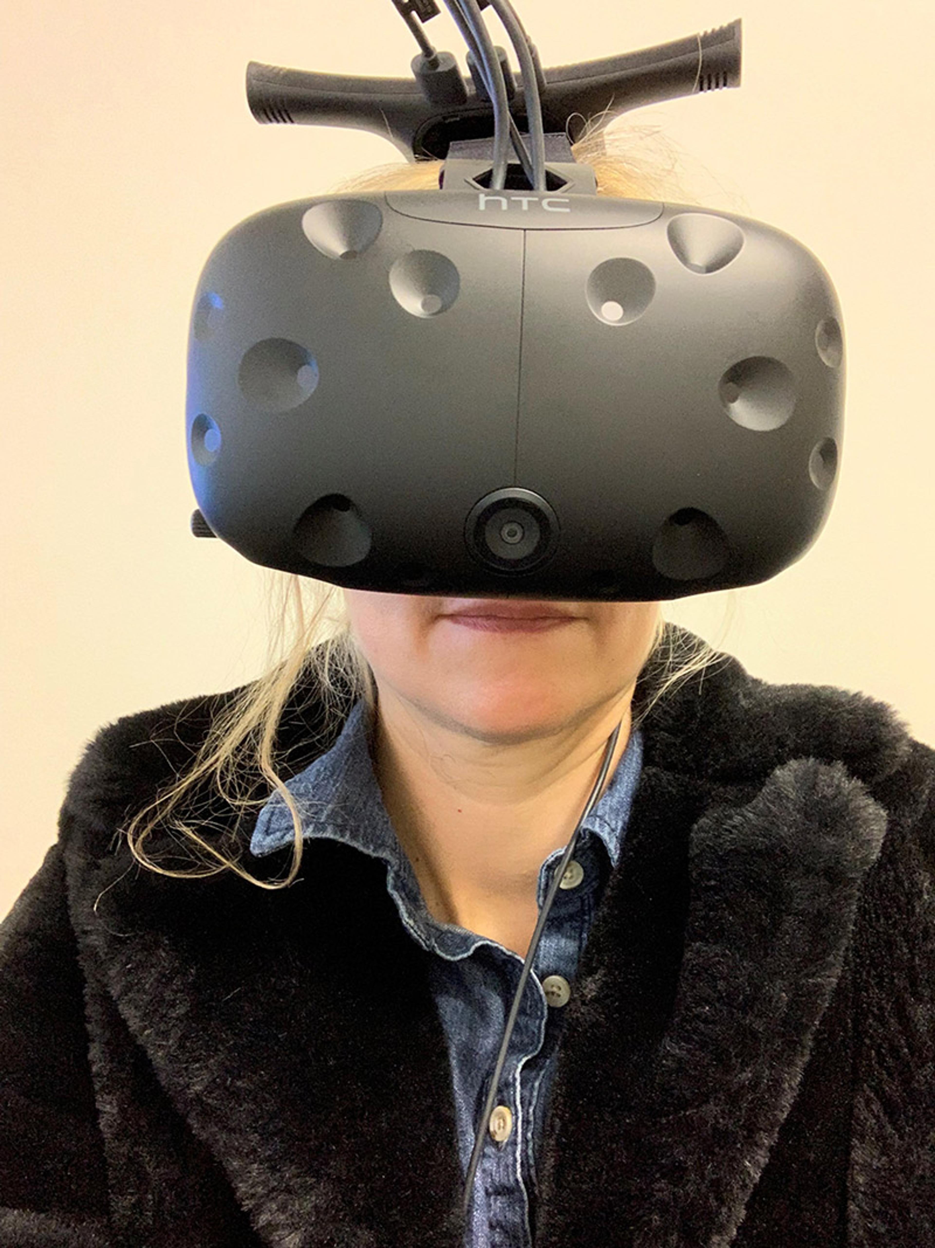 A woman wearing a virtual reality headset with cables attached, dressed in a denim shirt and a dark jacket, smiling slightly.