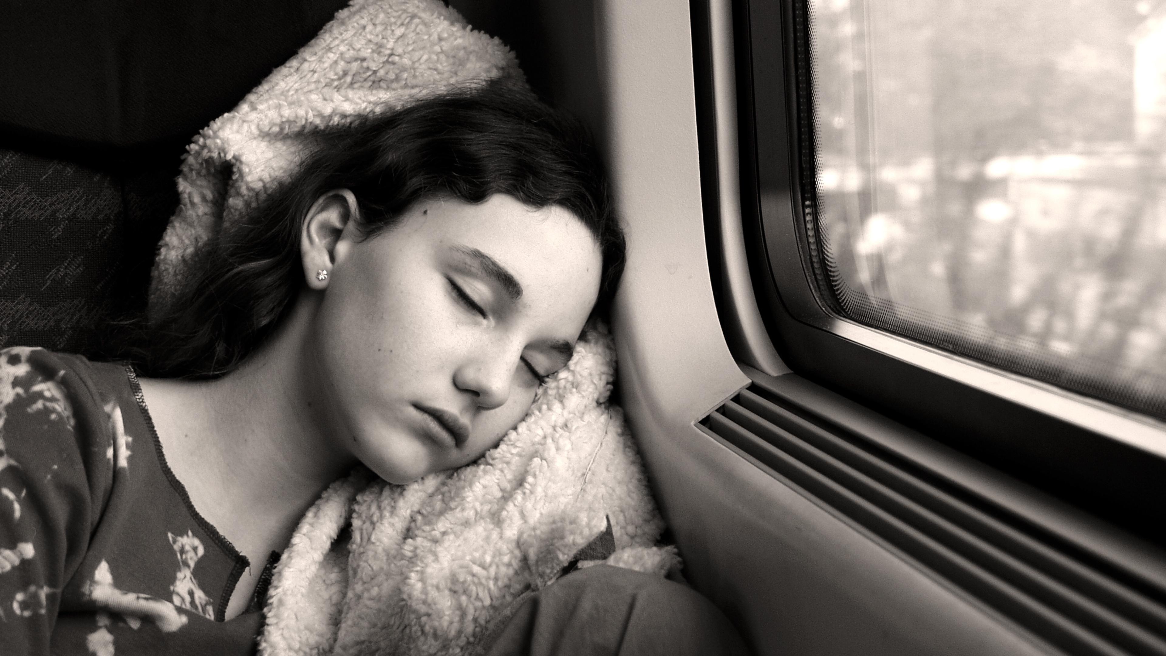 Black and white photo of a person sleeping on a train with their head on a blanket near a window with a blurred view outside.