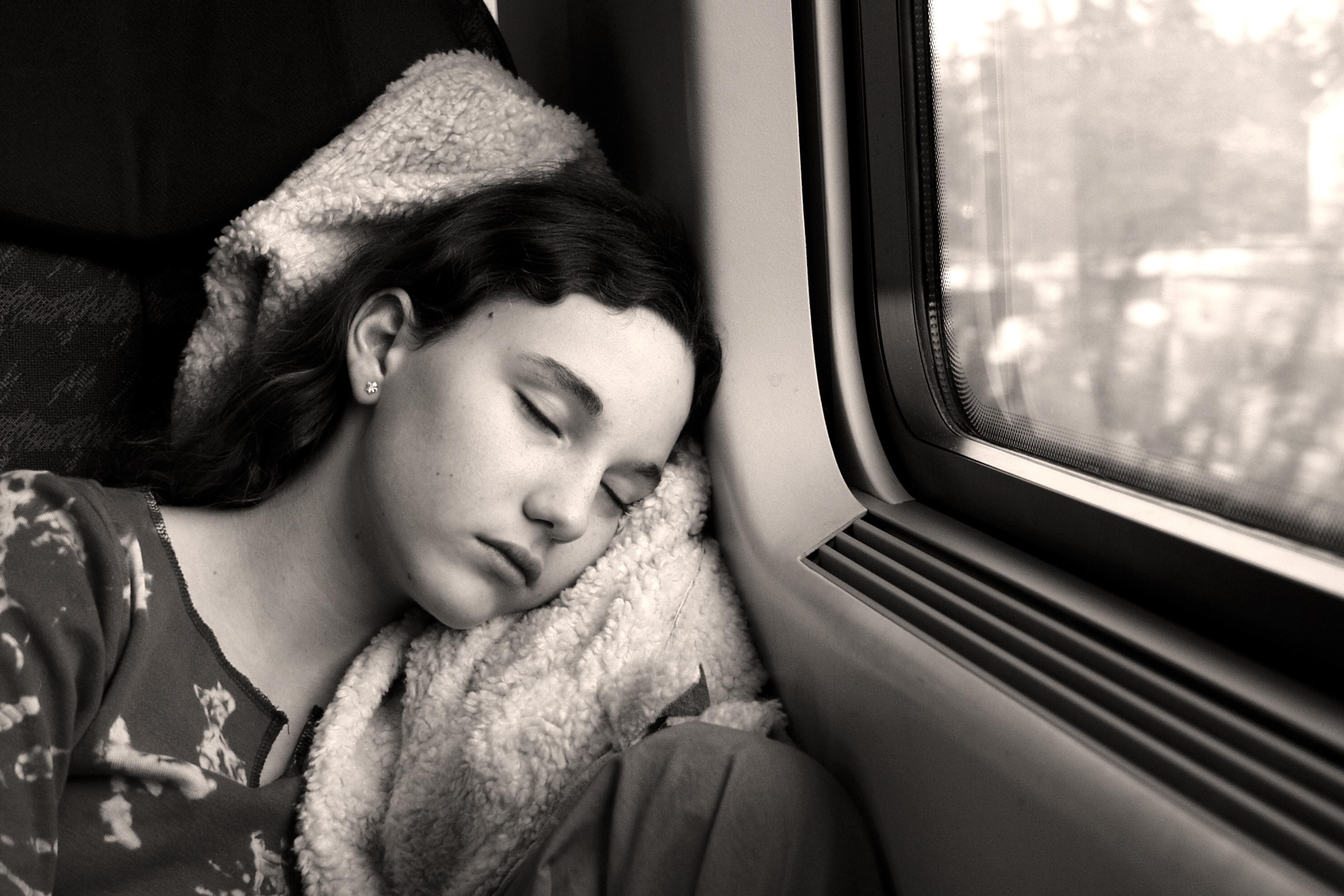 Black and white photo of a person sleeping on a train with their head on a blanket near a window with a blurred view outside.