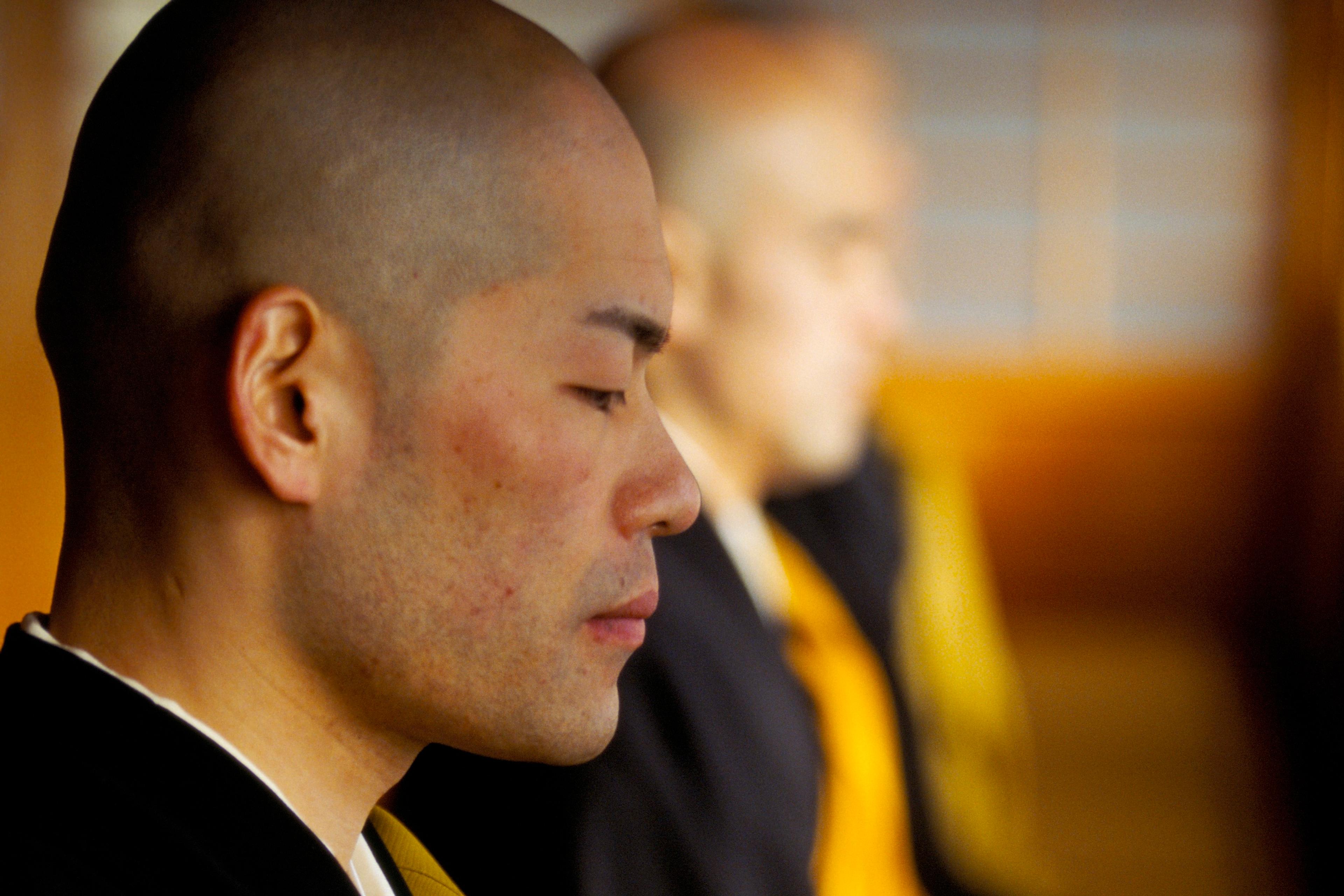 Photo of a meditating monk with closed eyes in a yellow and black robe, another monk blurred in the background.