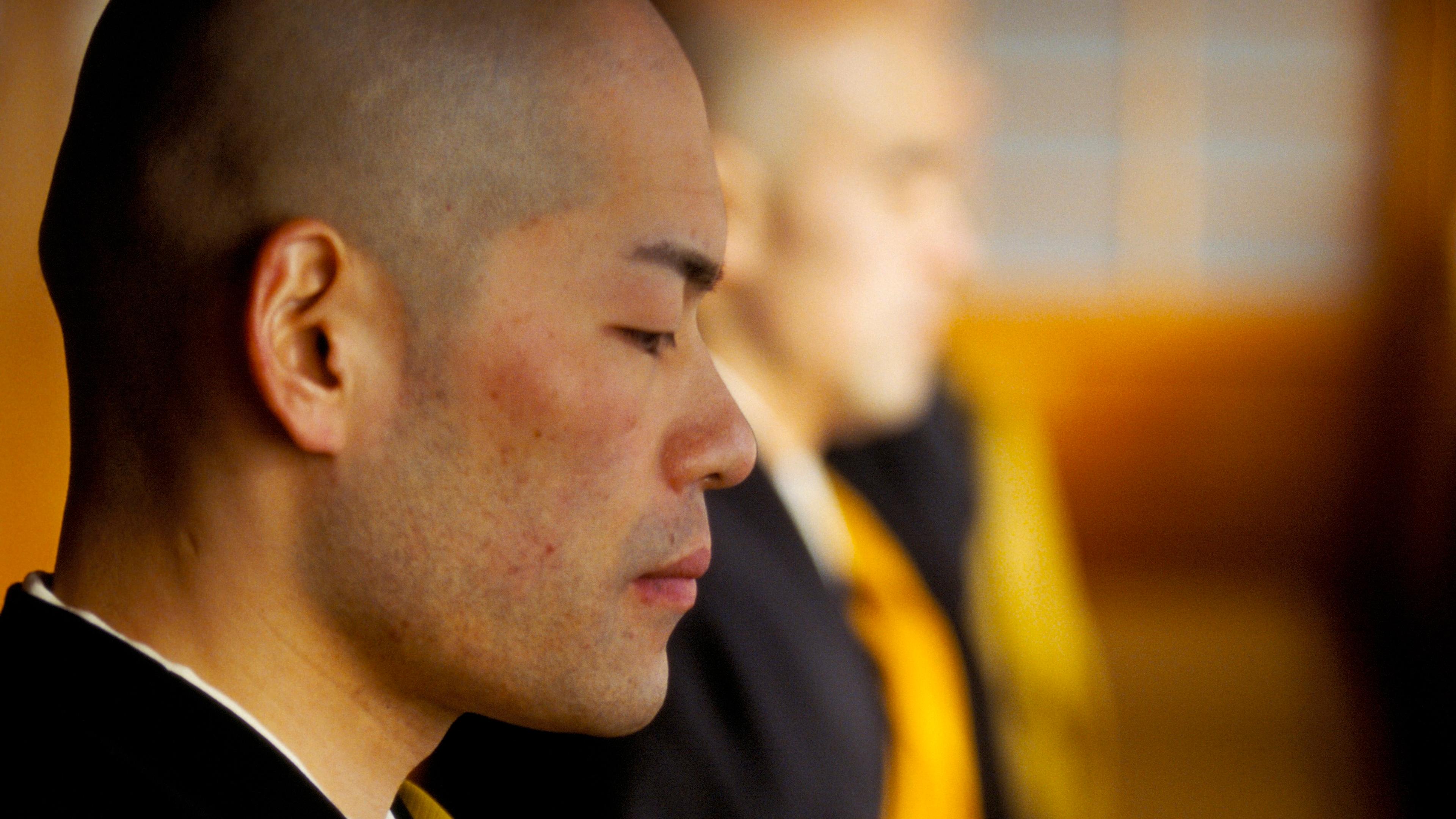 Photo of a meditating monk with closed eyes in a yellow and black robe, another monk blurred in the background.