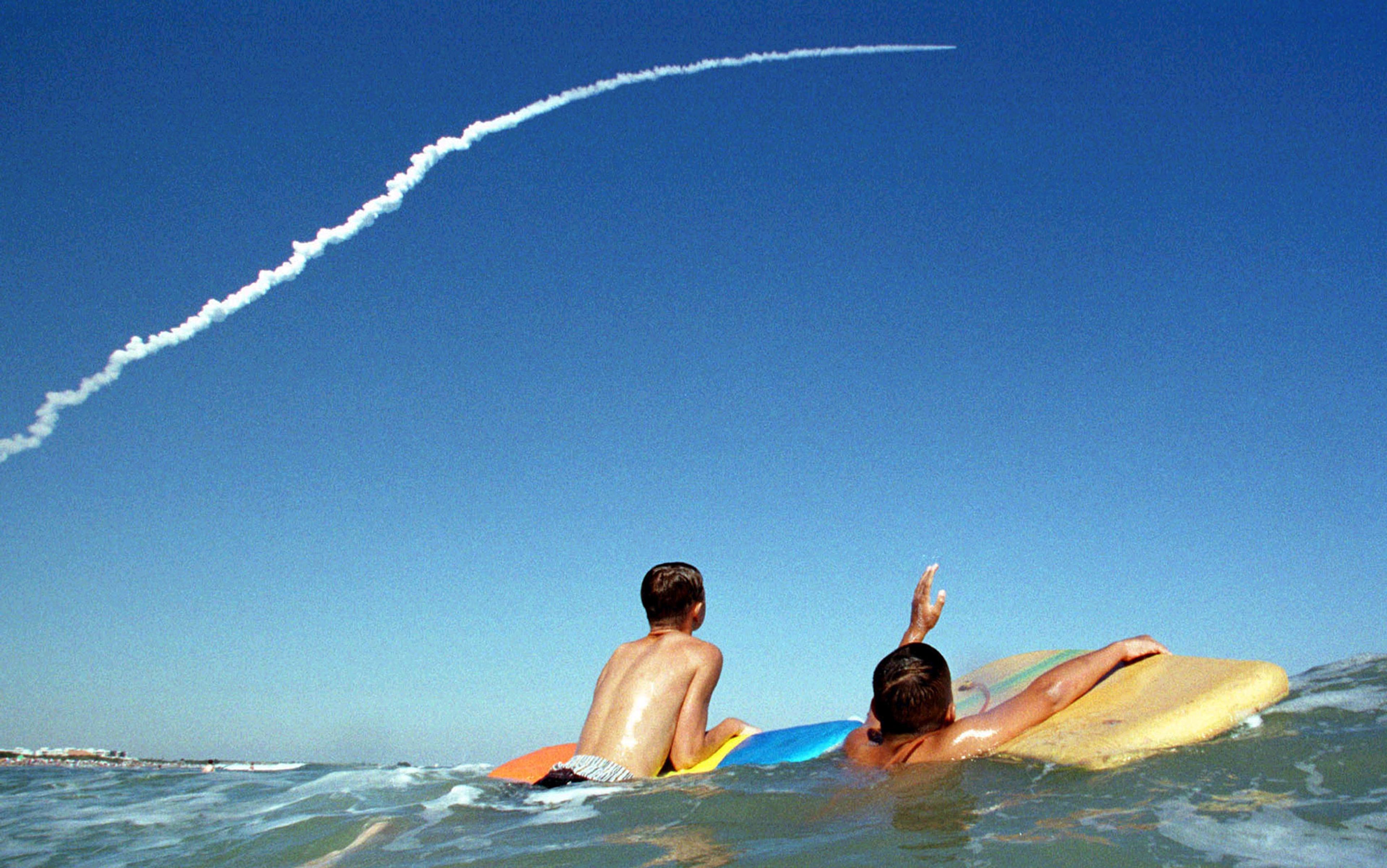 Photo of two boys on surfboards in the sea with one waving towards a rocket ascent leaving a trail in the clear blue sky