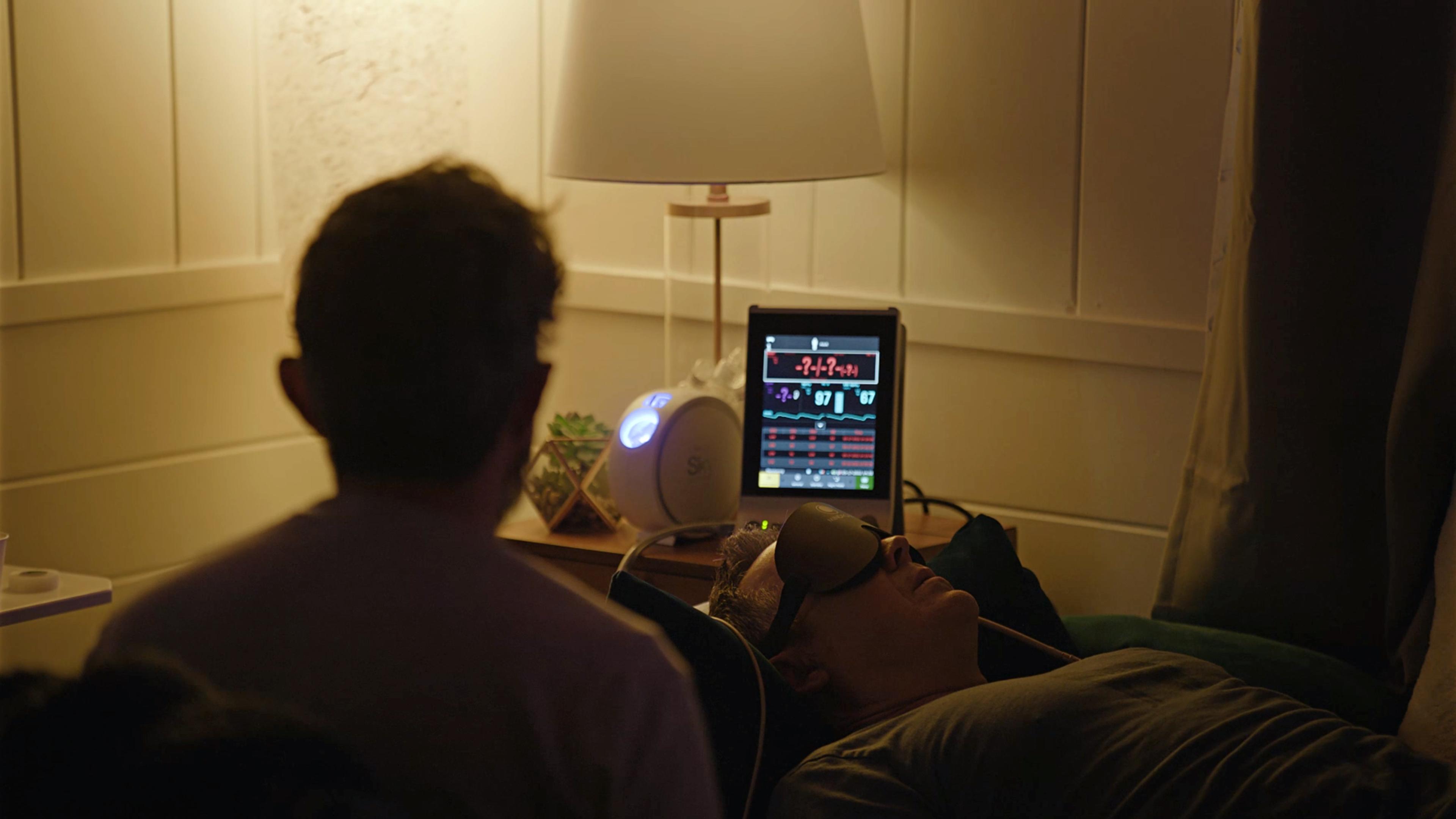 A man wearing an eye mask lies on a bed while another person observes; a monitor and lamp are on the nearby table.