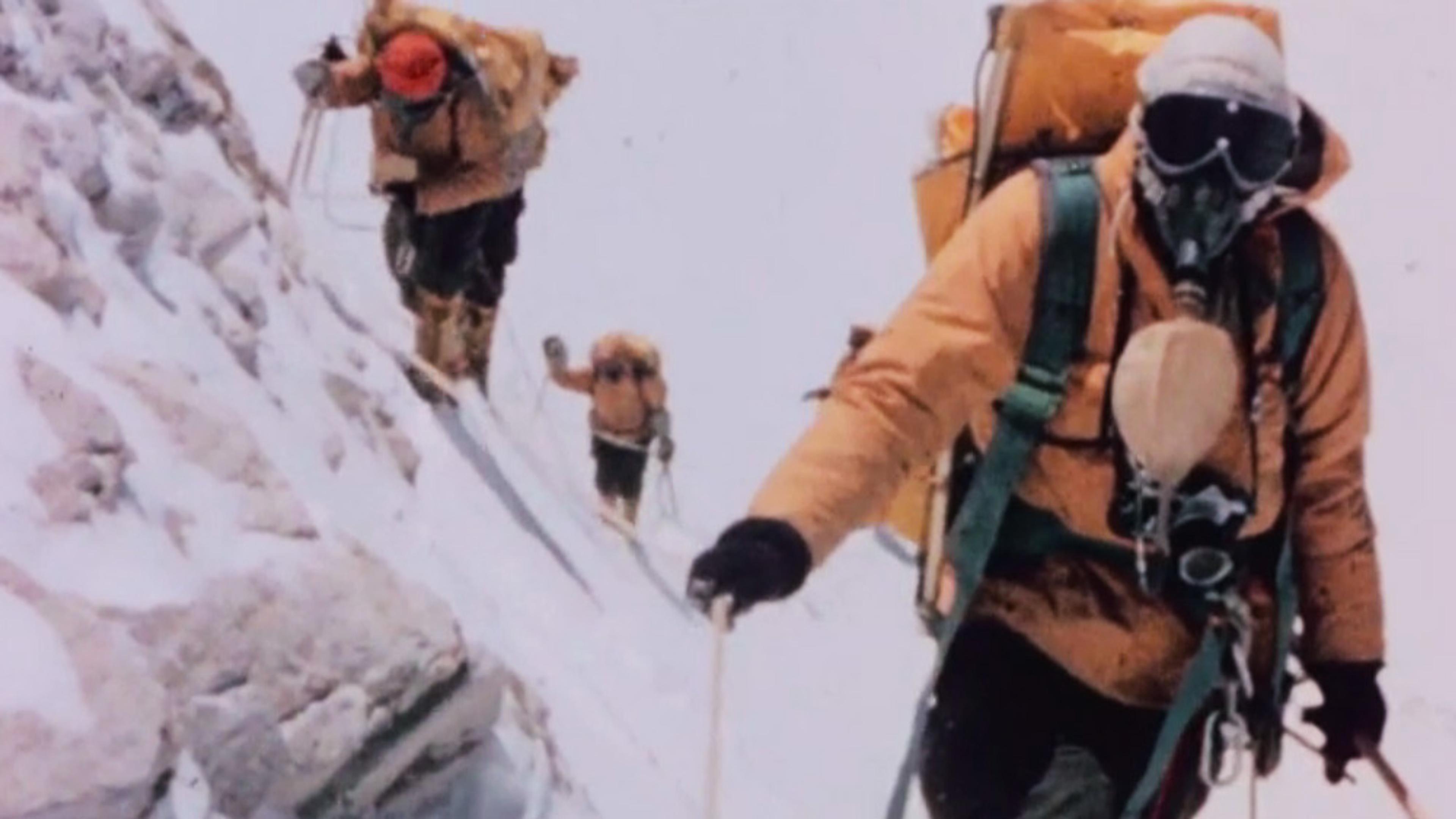 Three mountaineers in yellow jackets with oxygen masks and backpacks climbing a snowy mountain slope, using ropes.