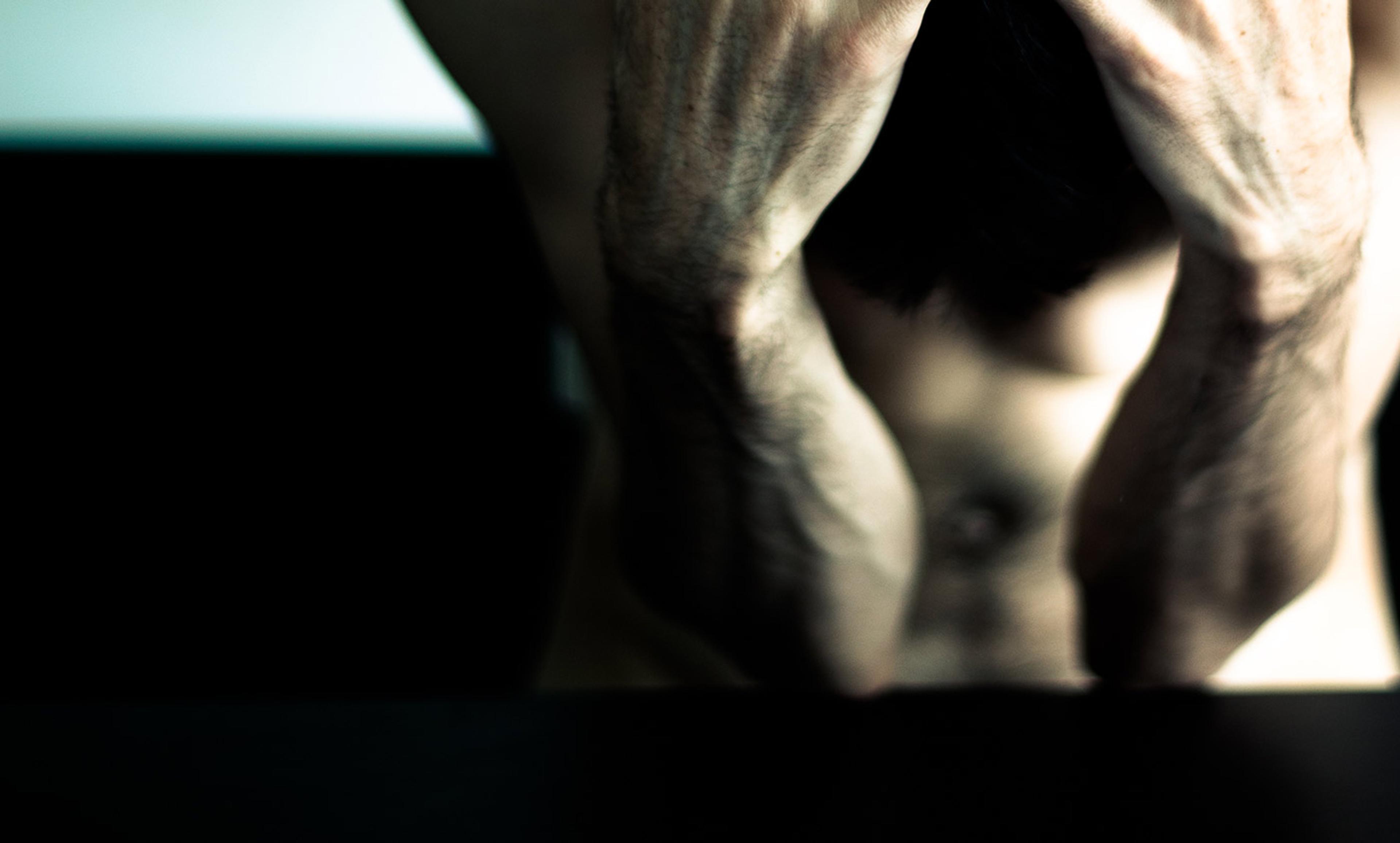 A shirtless person with their head in their hands, arms and back muscles are visible; dim lighting and high contrast.