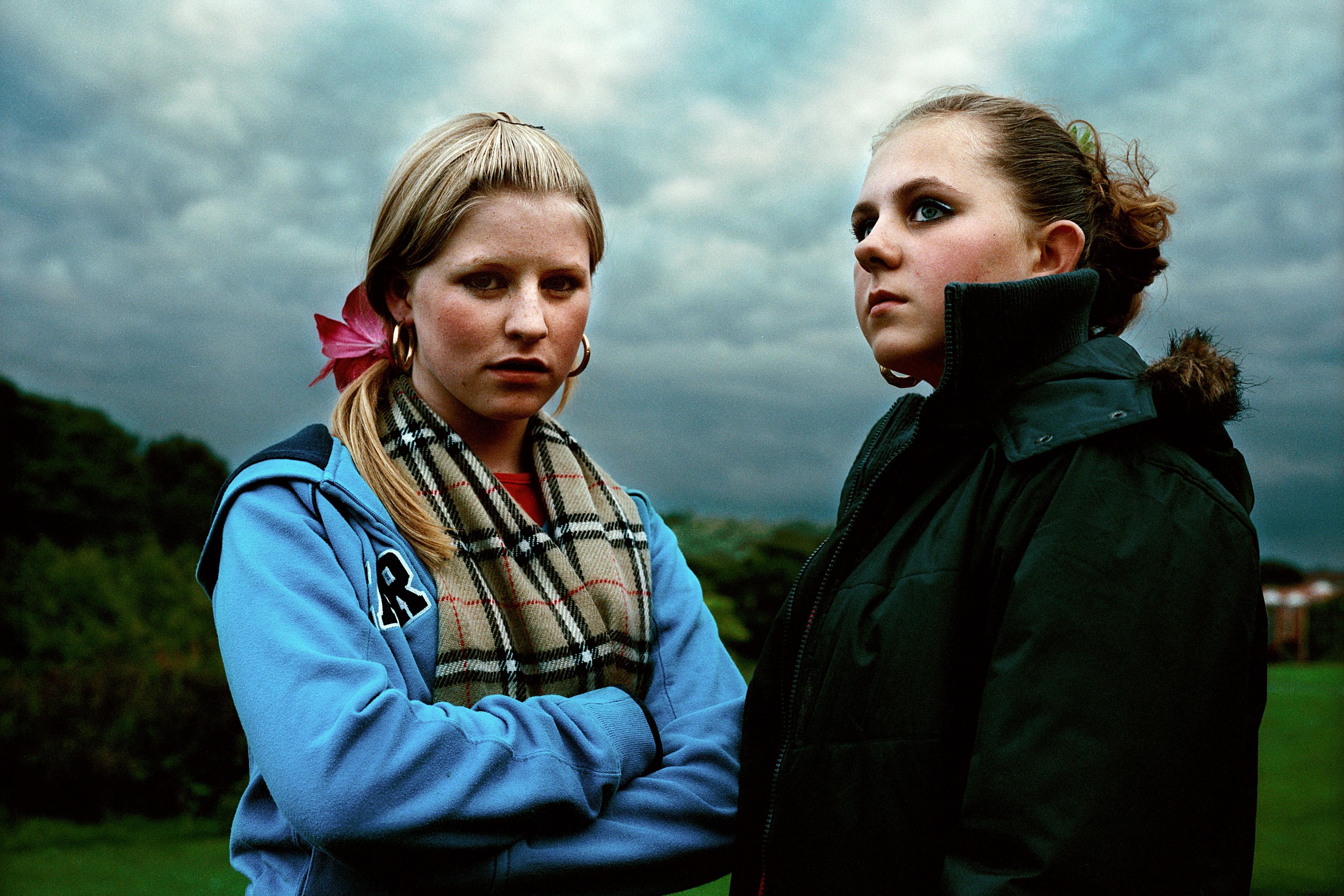 Photo of two young women standing outdoors with cloudy sky, one in a blue hoodie and scarf the other in a dark coat.