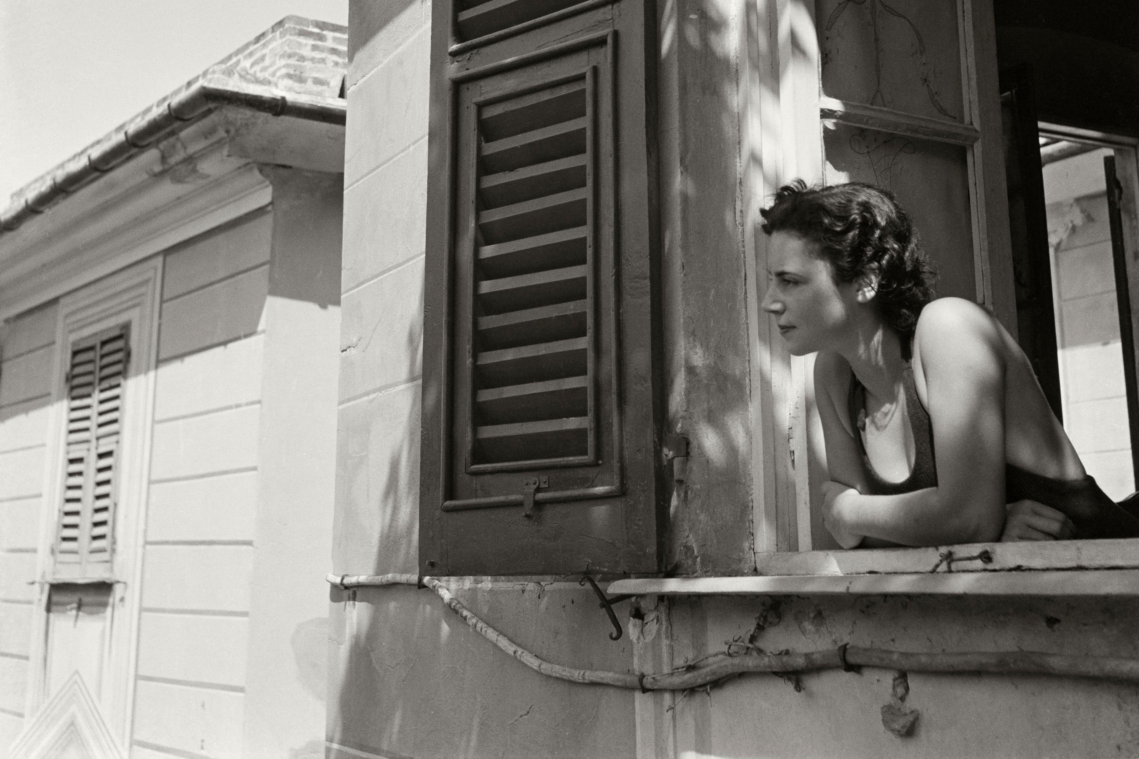 Black and white photo of a woman leaning out of a window in a building with closed shutters.