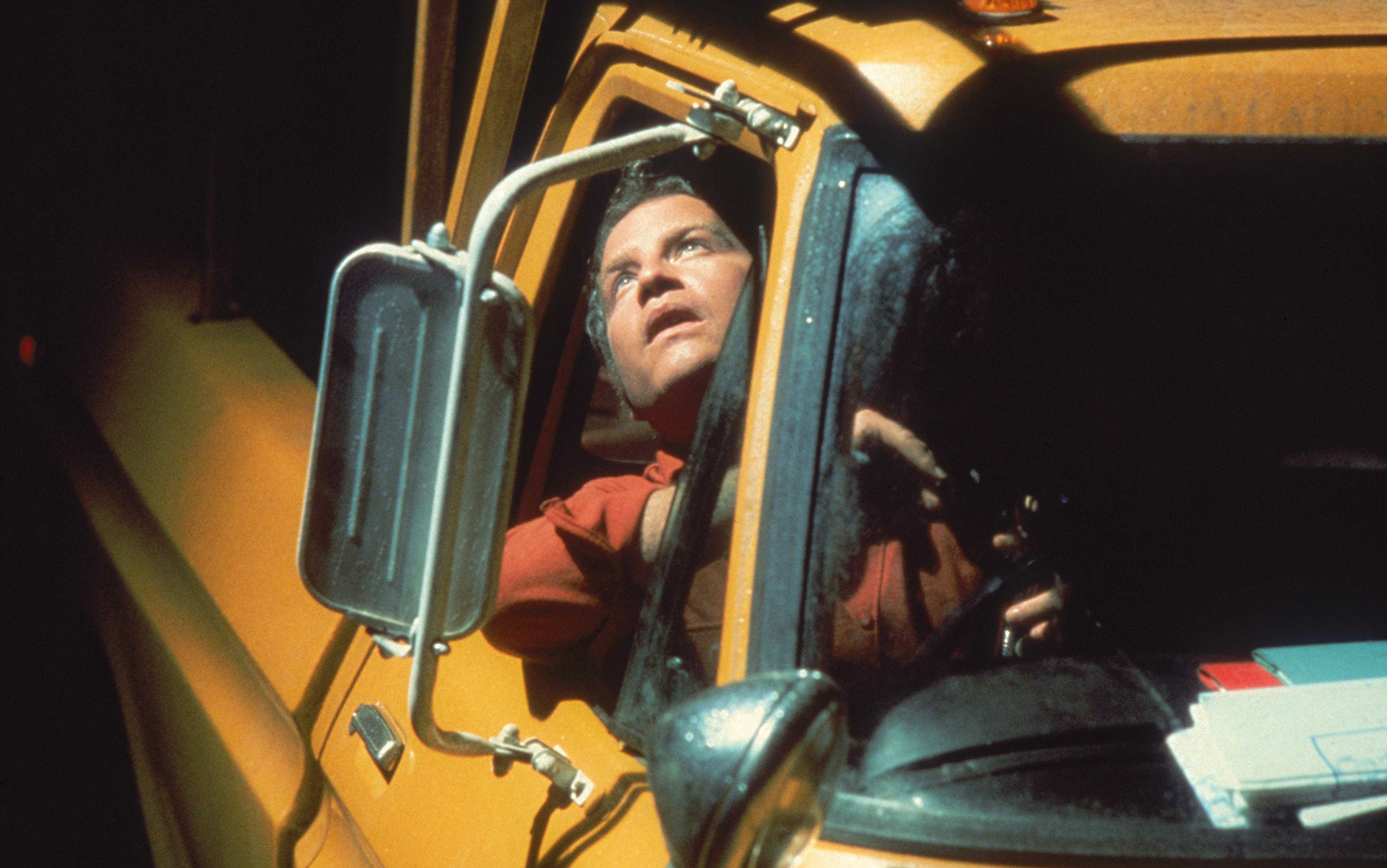A man looking up from the driver’s seat of a yellow vehicle with an open window and side mirror visible.