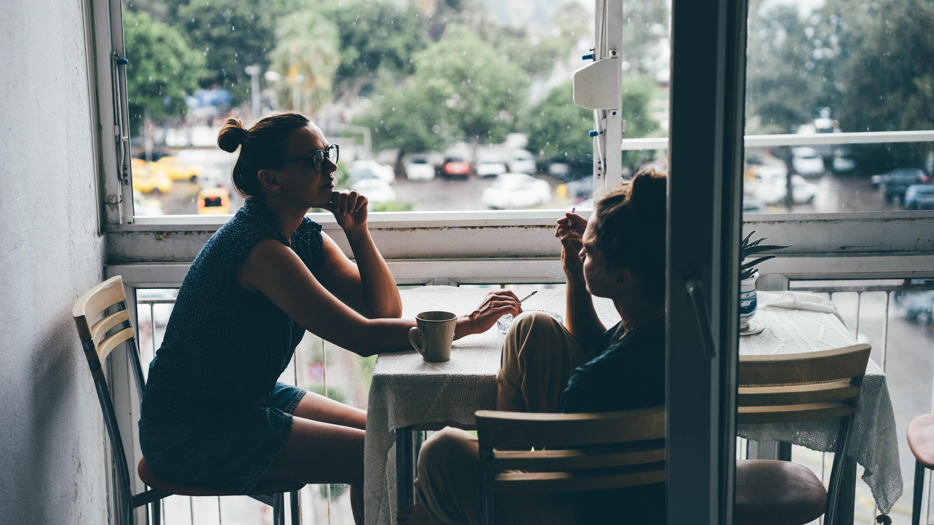 Photo of two people sitting at a table by a window, talking and drinking coffee, with a rainy view outside.
