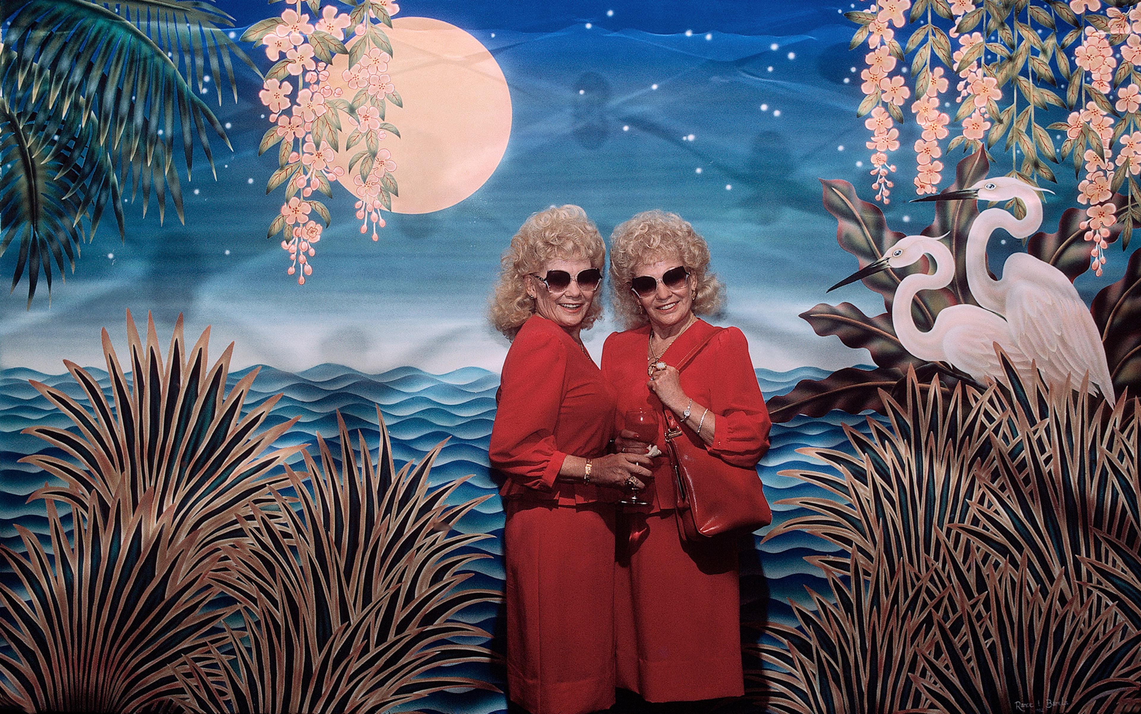 Two elderly women in red dresses and sunglasses standing in front of a tropical mural with birds, flowers, and a full moon.