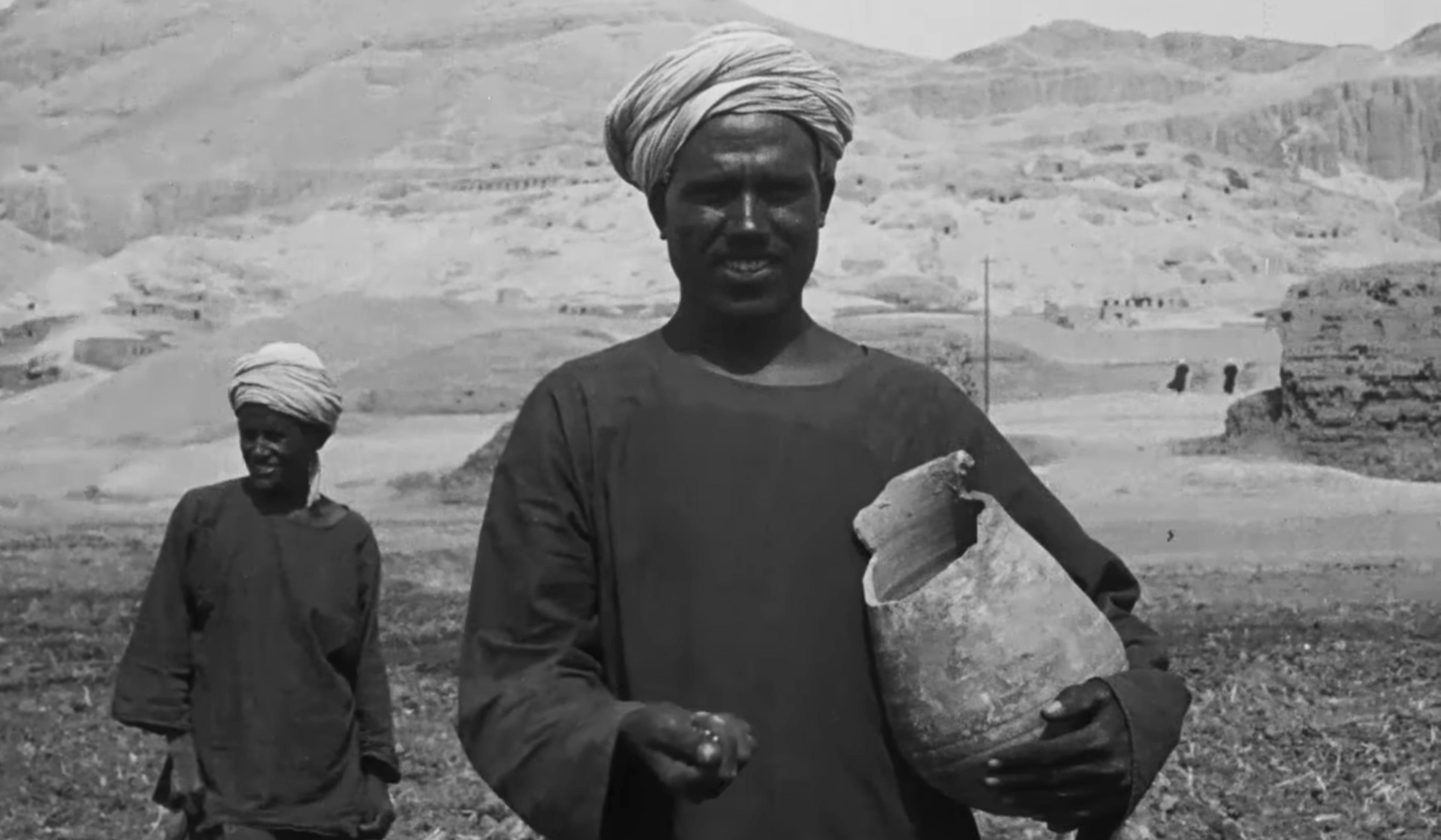 Black-and-white photo of two Egyptian men in traditional attire, one holding a broken pot, standing in a desert landscape with mountains.