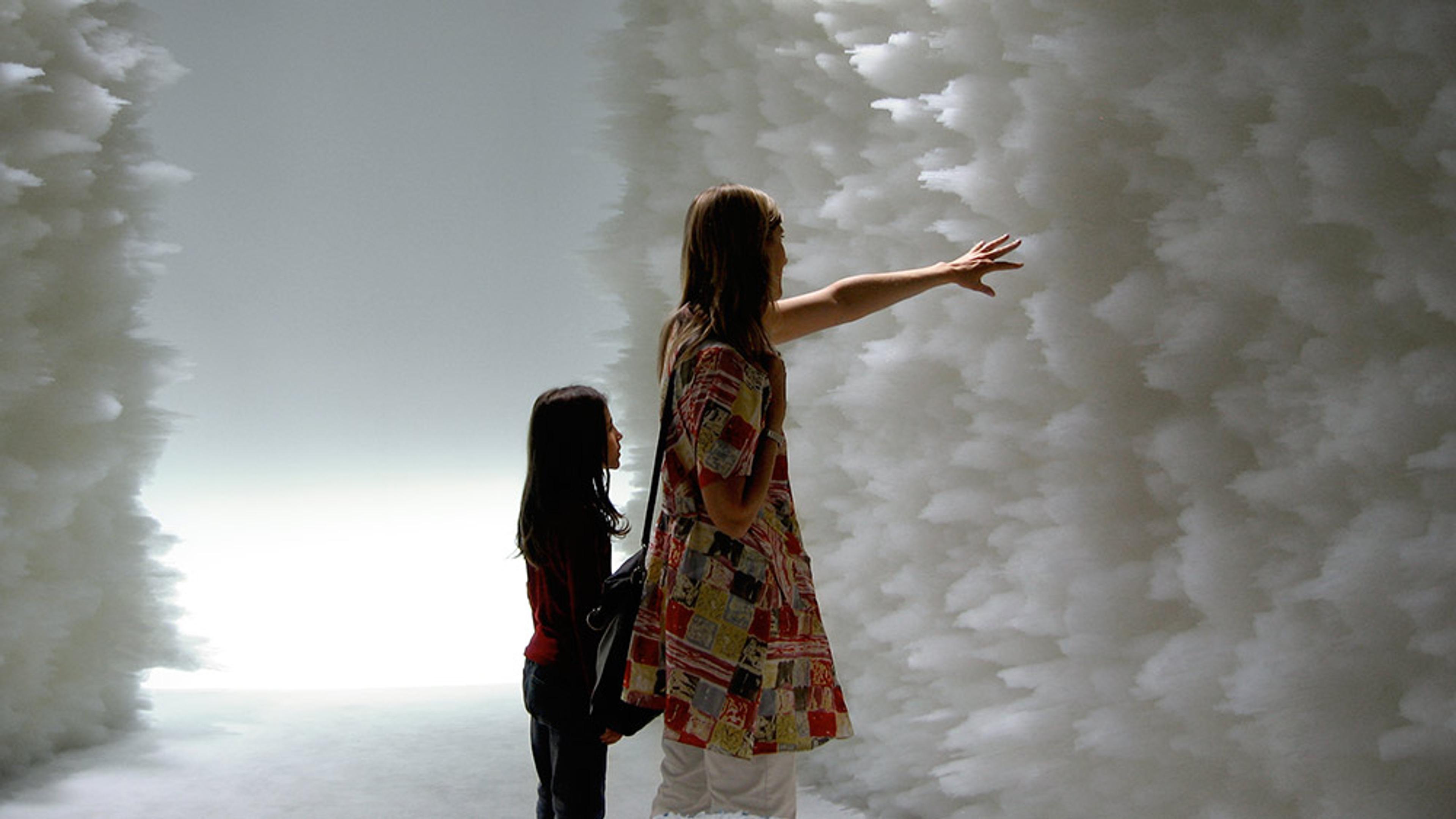 A woman and child standing between two large walls covered in a fluffy, cloud-like texture, with the woman touching the surface.