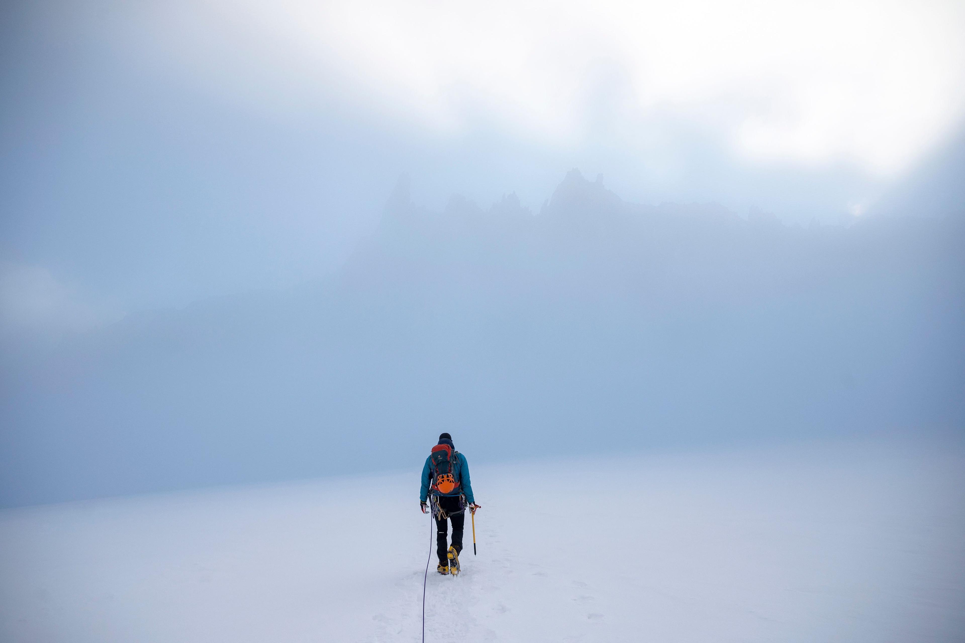 Photo of a lone mountaineer walking in misty snowy terrain with climbing gear and an ice axe, heading towards foggy peaks.