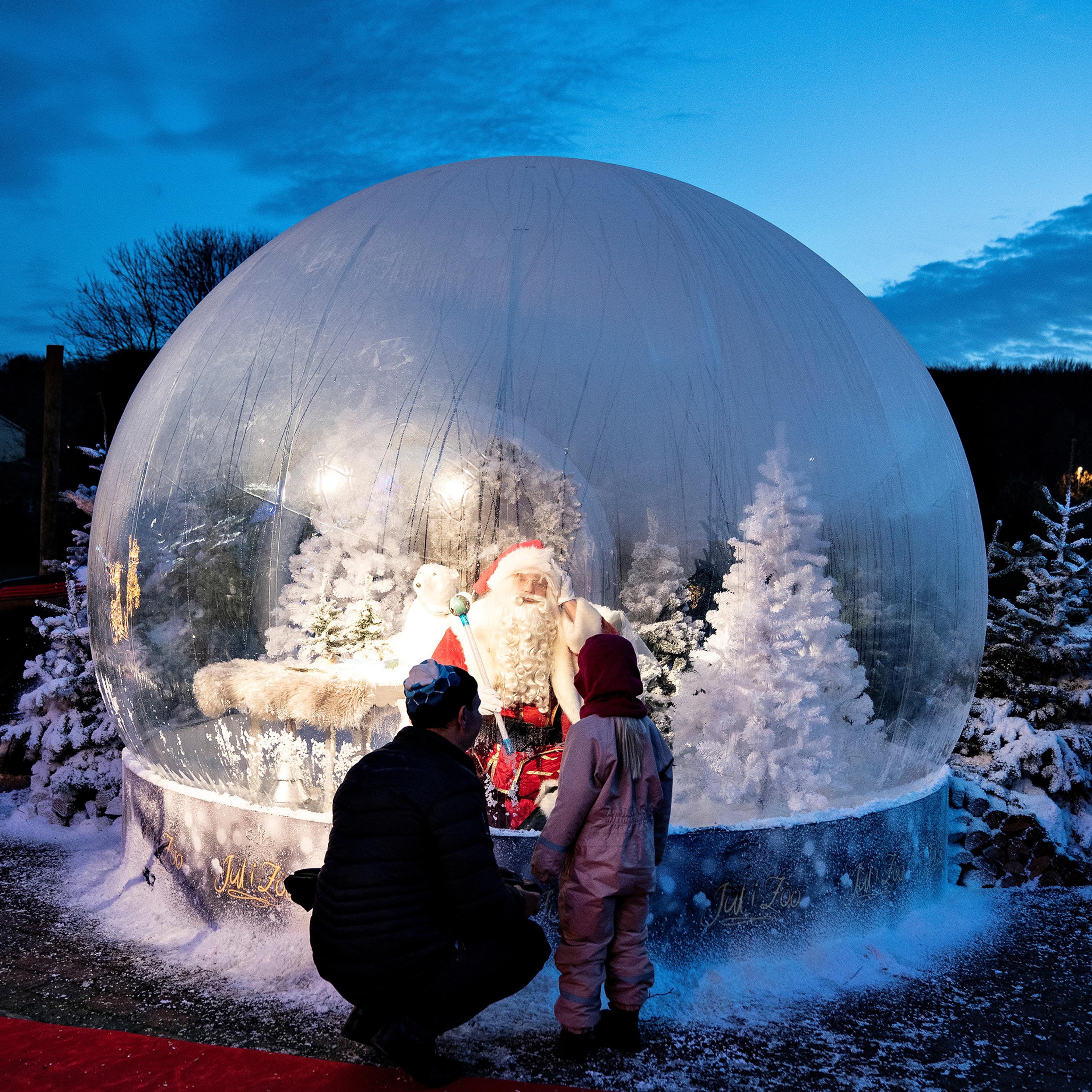 An adult and child looking at Santa inside a large snow globe with snowy trees at dusk.