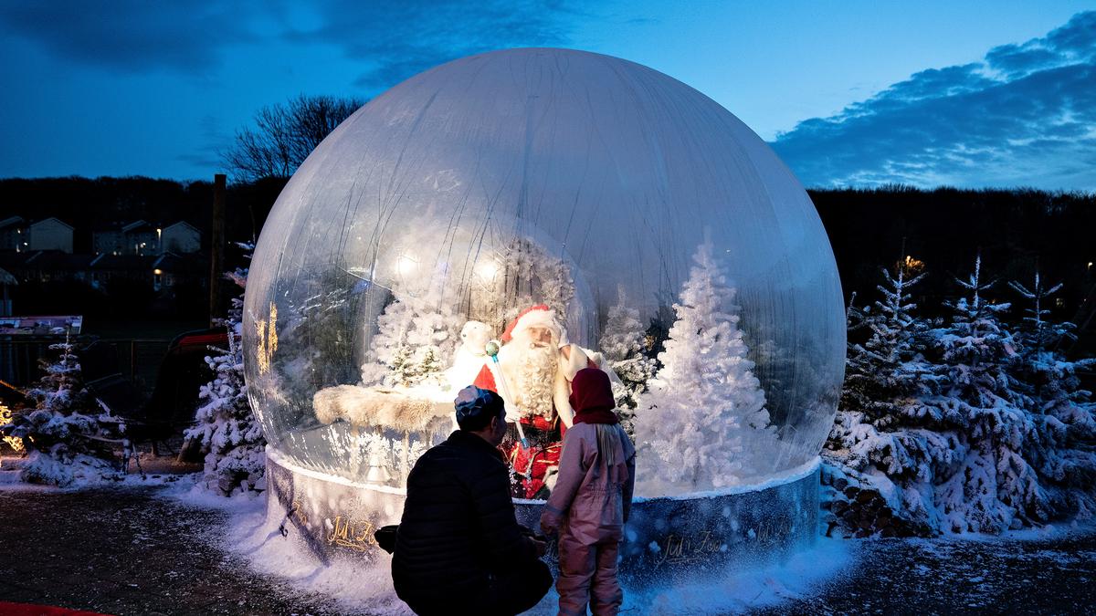 An adult and child looking at Santa inside a large snow globe with snowy trees at dusk.