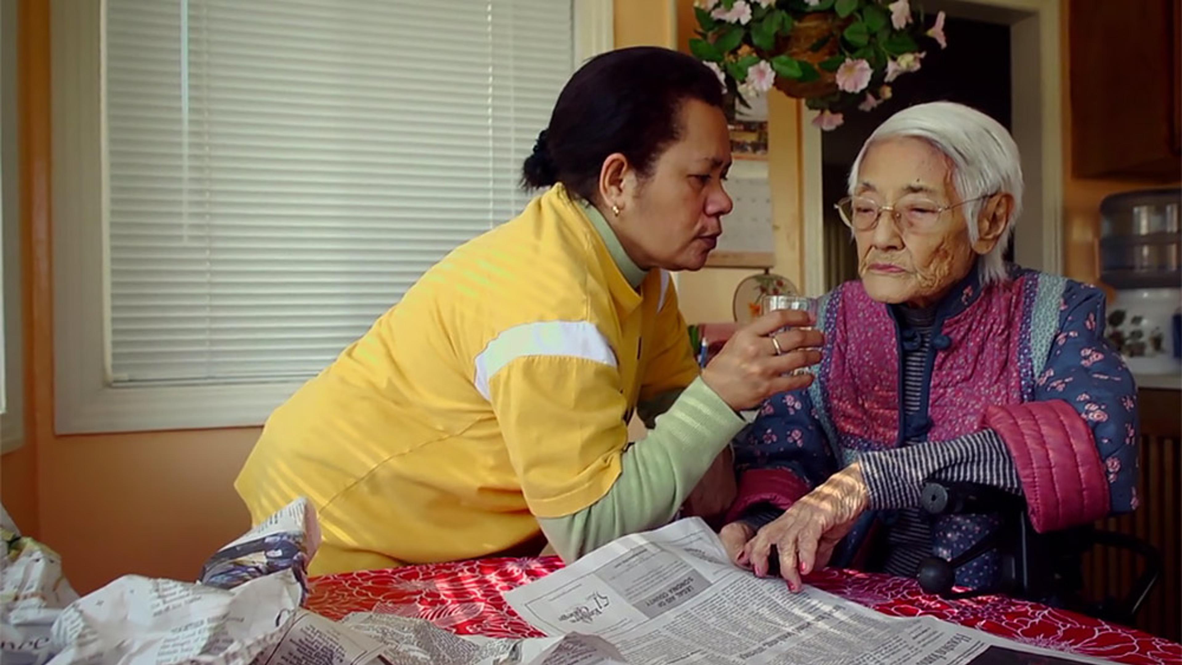A caregiver in a yellow shirt helping an elderly woman in glasses and a pink jacket to drink water at a kitchen table.