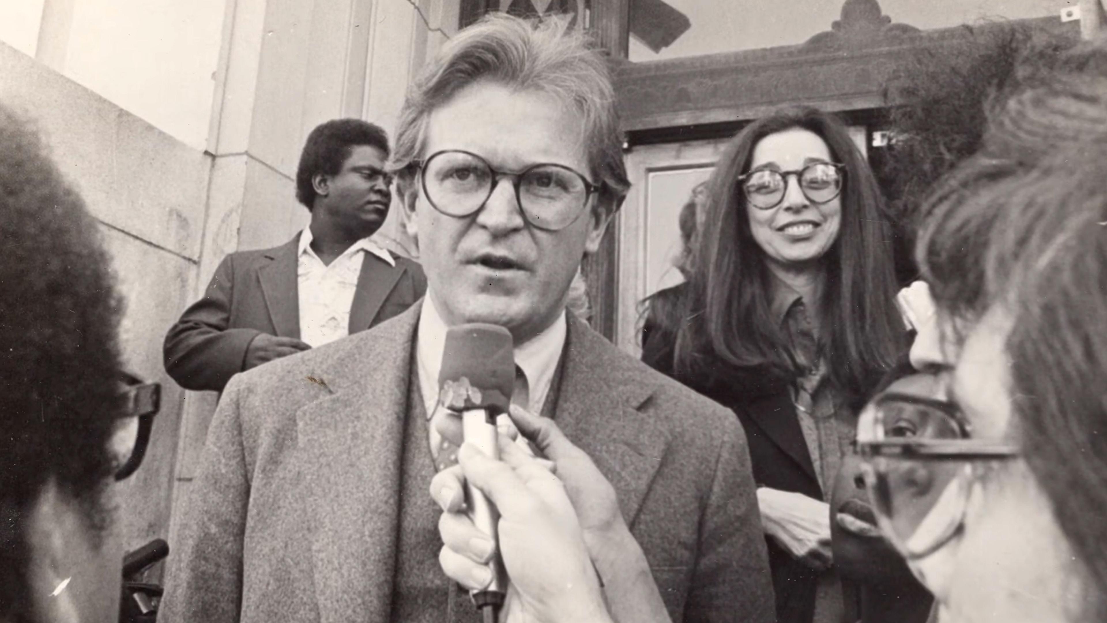 Black-and-white photo of a man speaking into microphones held by the press outside a building, with a woman smiling at him in the background.