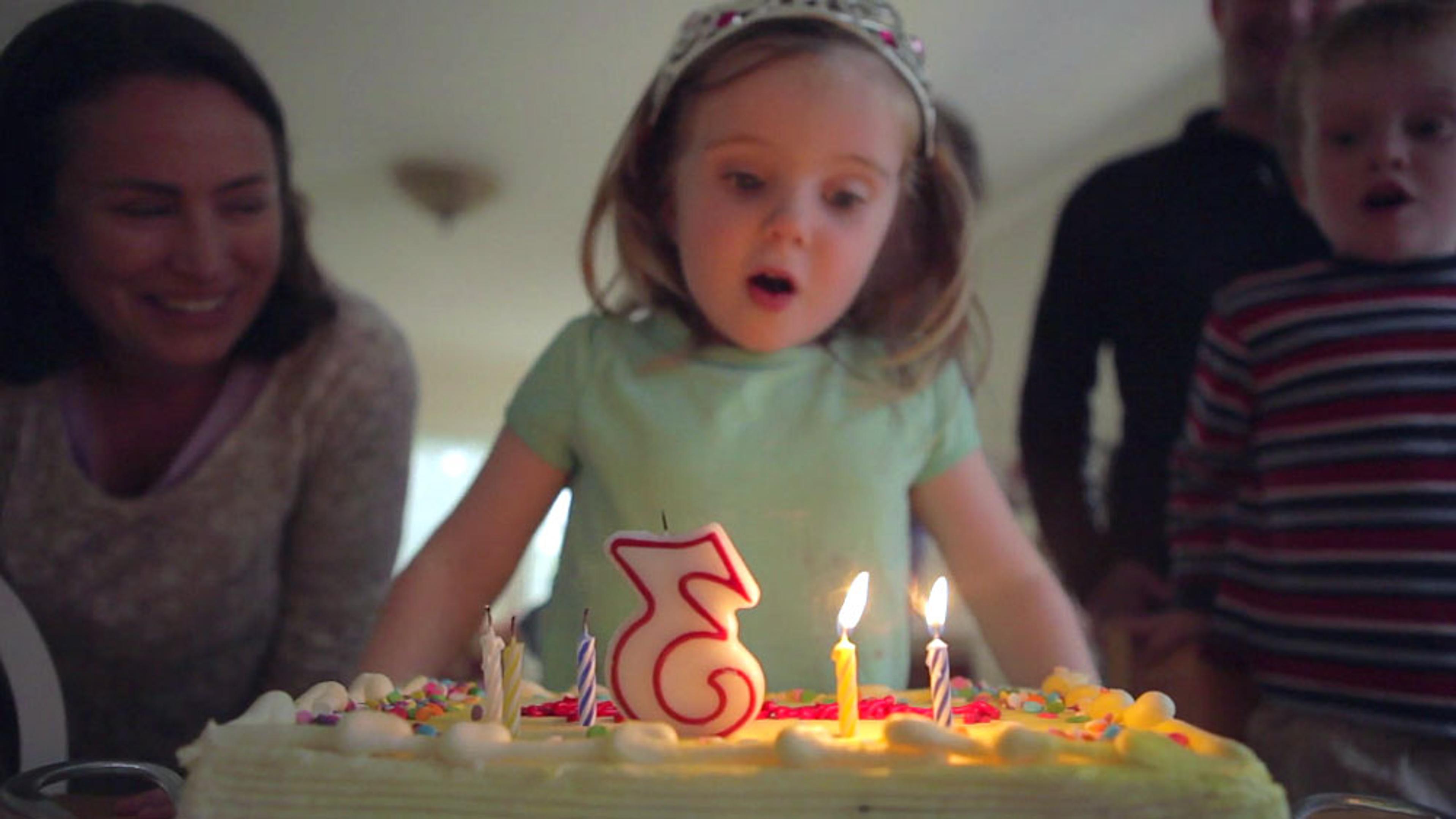 A young girl blowing out birthday candles on a cake with the number three, surrounded by family members.