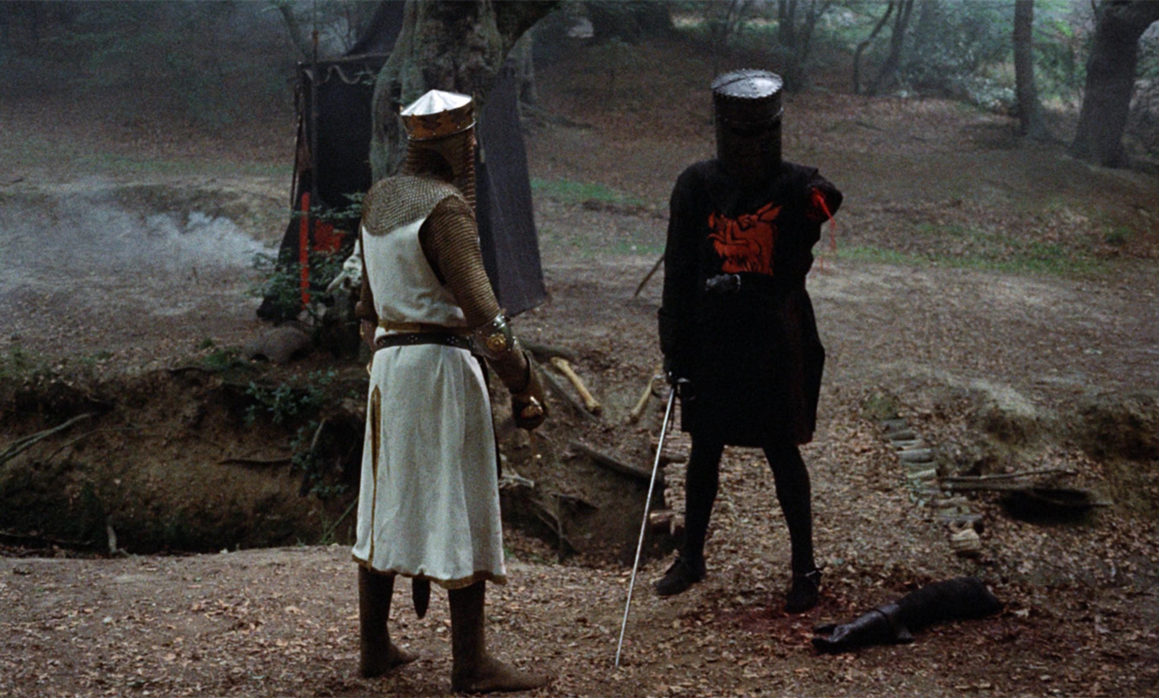 Film still with two medieval knights in armour standing in a forest, one missing an arm, with a tent in the background.