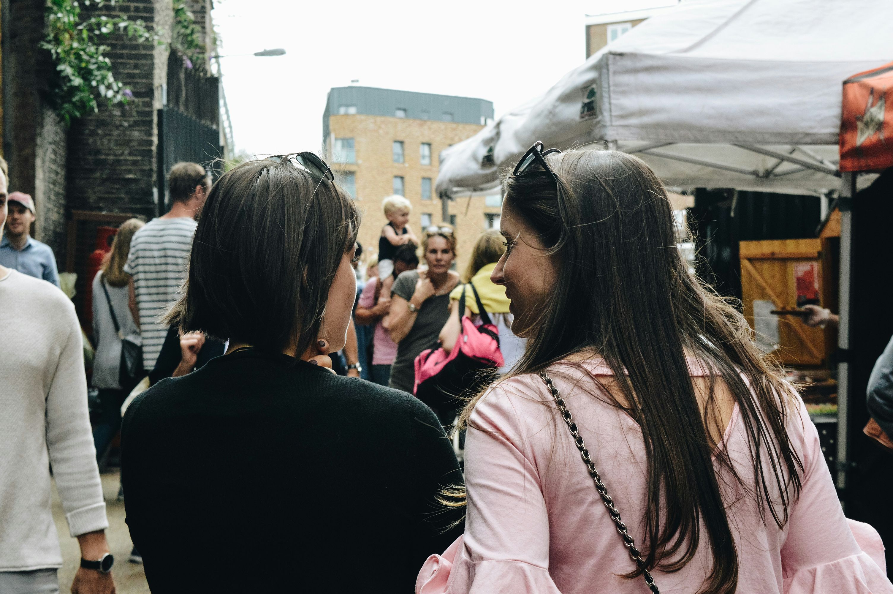 Two women chatting at an outdoor market with stalls and people in the background on a cloudy day.