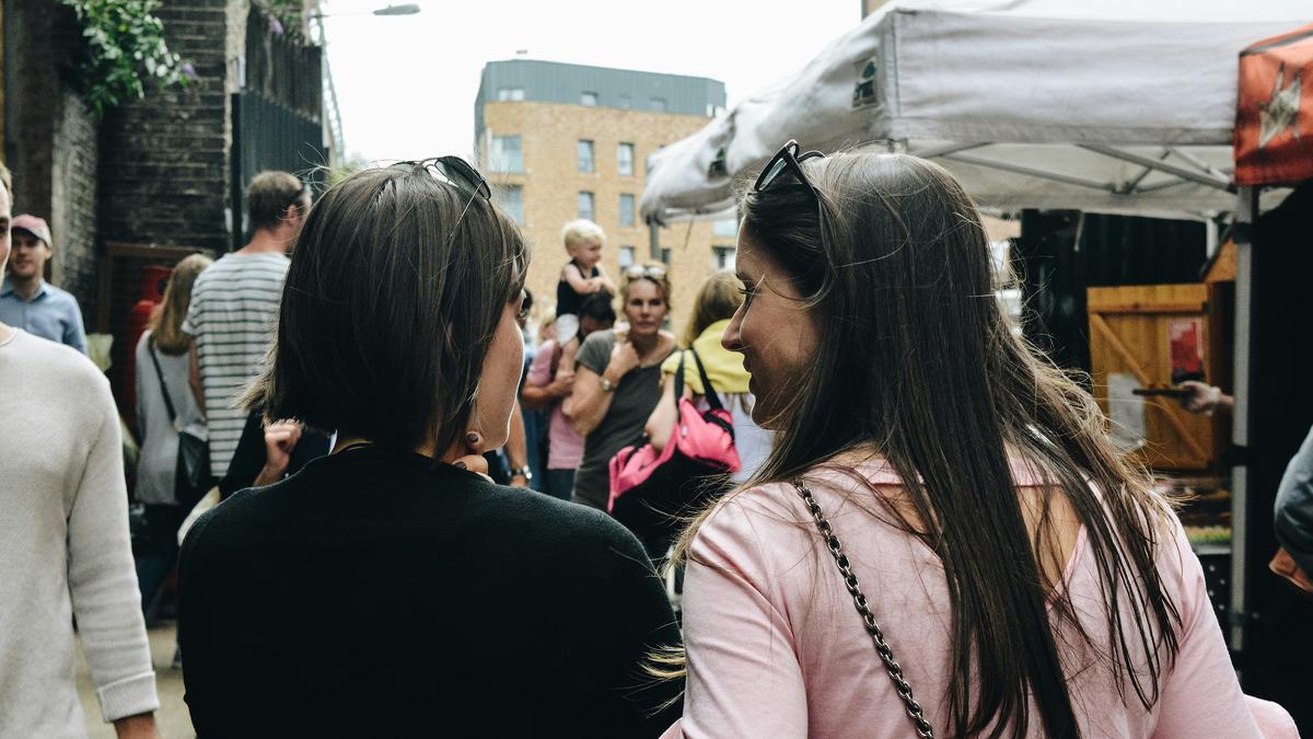 Two women chatting at an outdoor market with stalls and people in the background on a cloudy day.