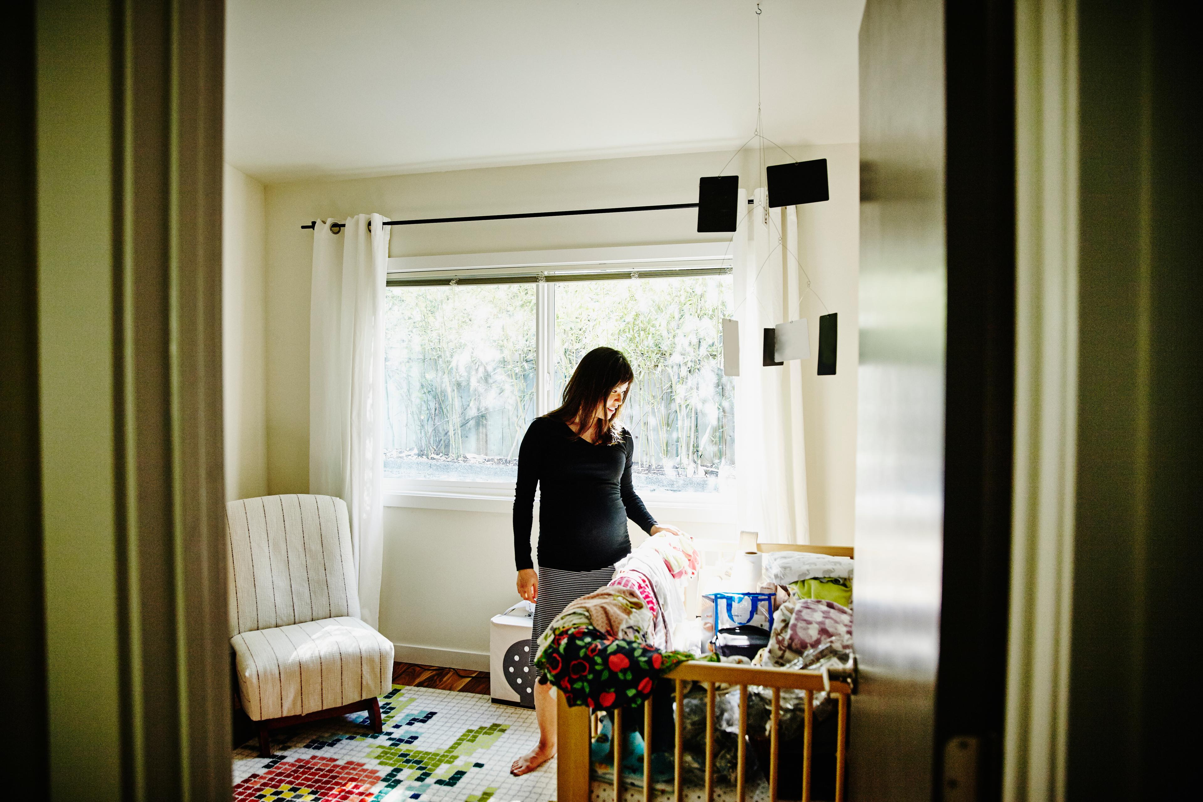 Photo of a woman in a nursery next to a crib with colourful blankets and a chair by a window with white curtains.