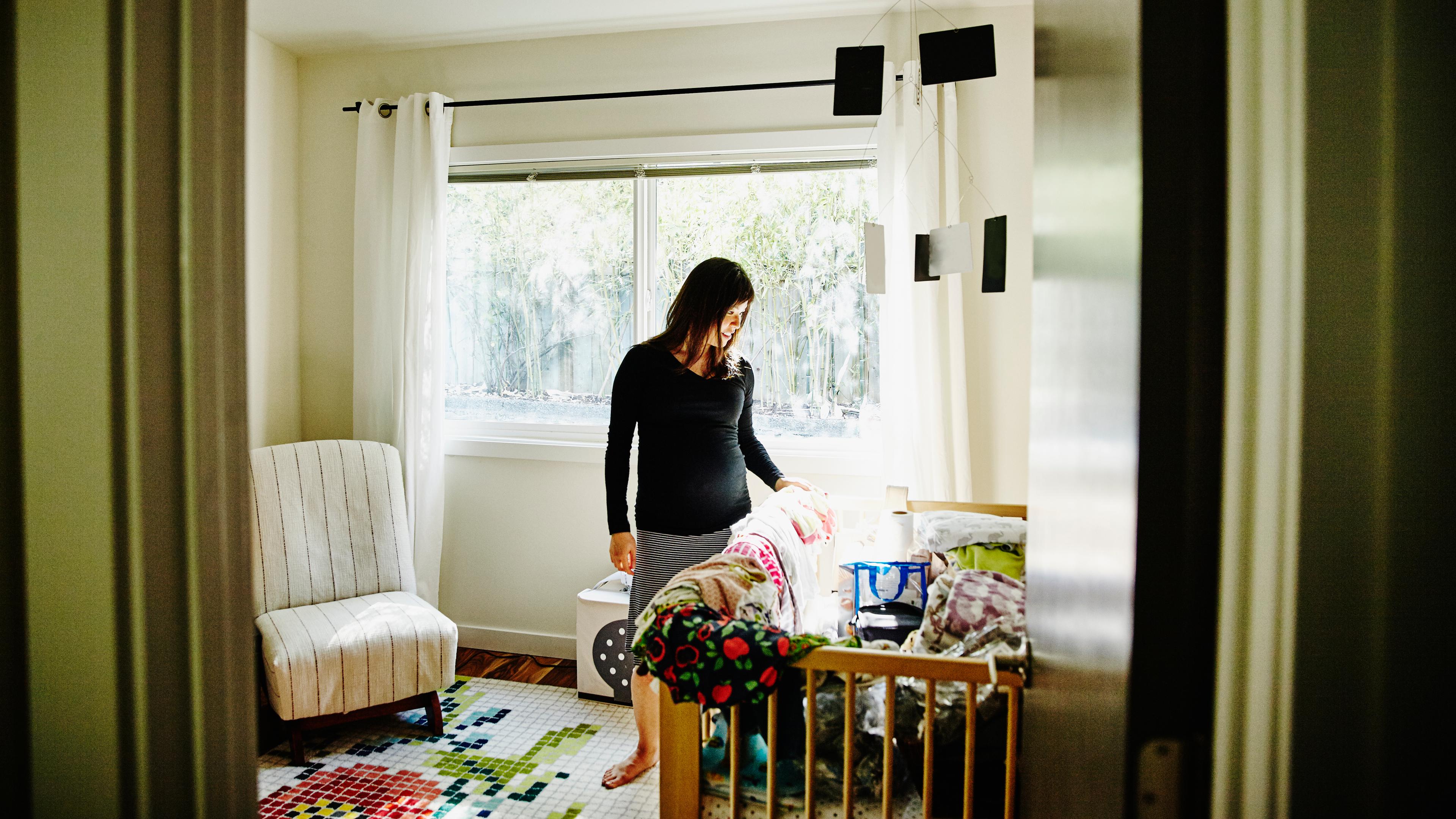 Photo of a woman in a nursery next to a crib with colourful blankets and a chair by a window with white curtains.