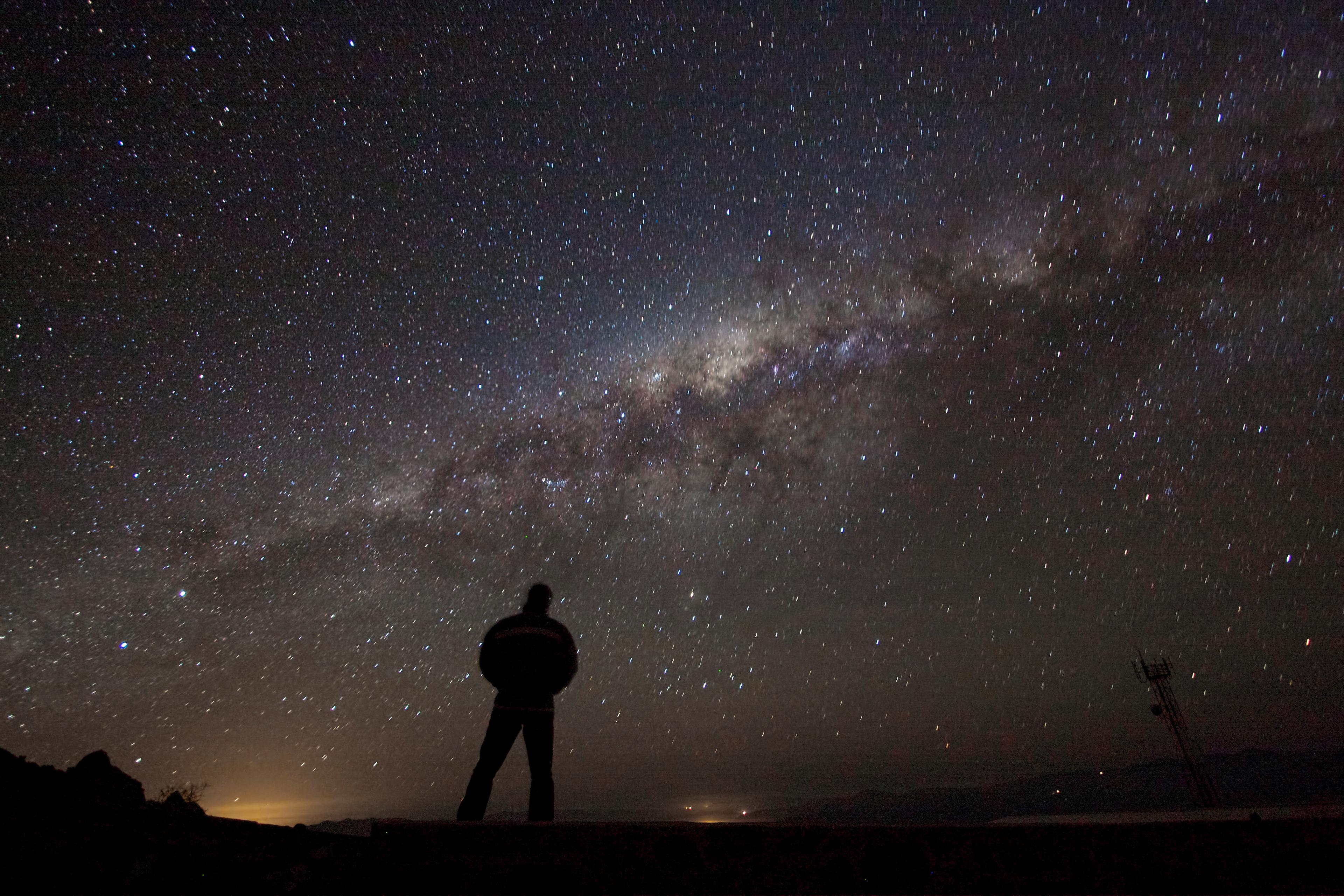 Photo of a person silhouetted against the Milky Way galaxy in a starry night sky, with a faint horizon glow.