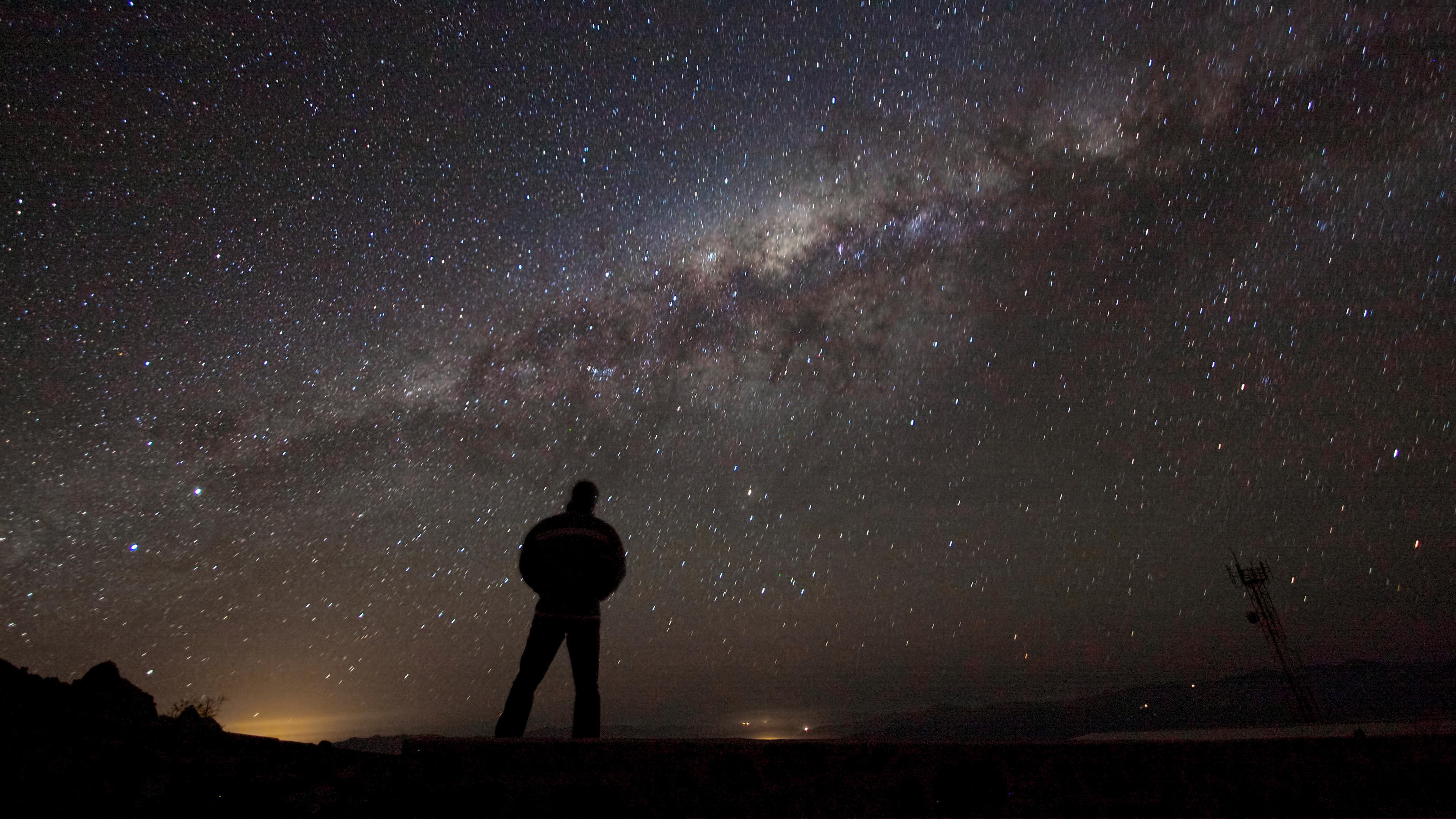 Photo of a person silhouetted against the Milky Way galaxy in a starry night sky, with a faint horizon glow.