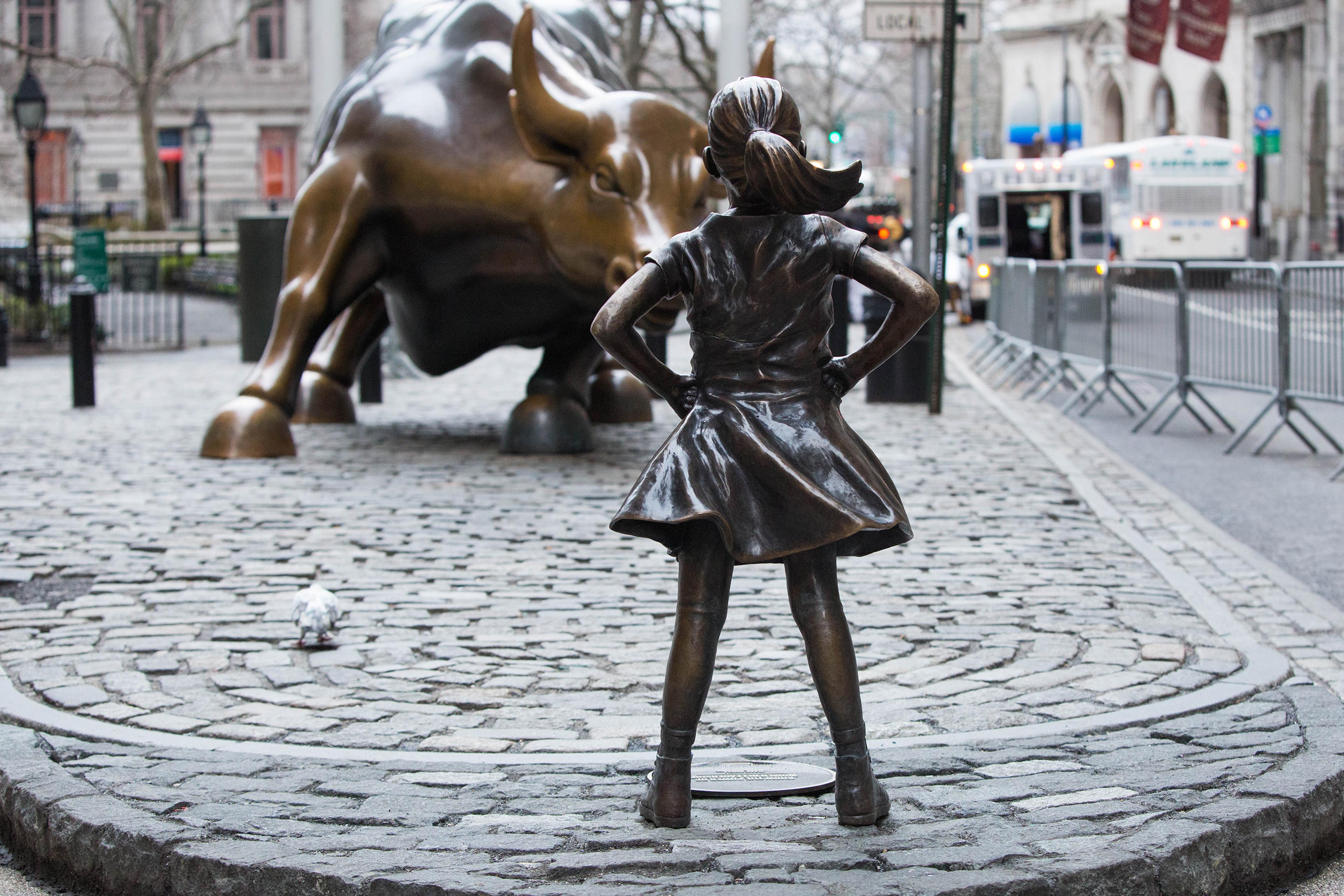 A bronze statue of a girl facing a large bull sculpture on a cobblestone street in an urban setting.