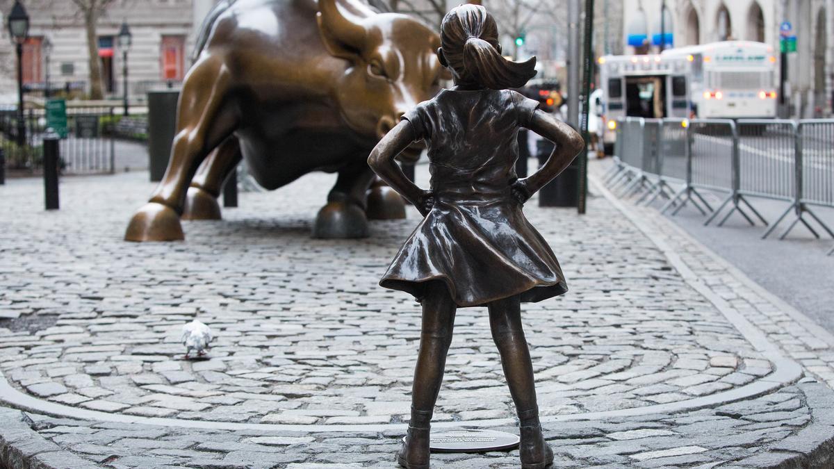 A bronze statue of a girl facing a large bull sculpture on a cobblestone street in an urban setting.