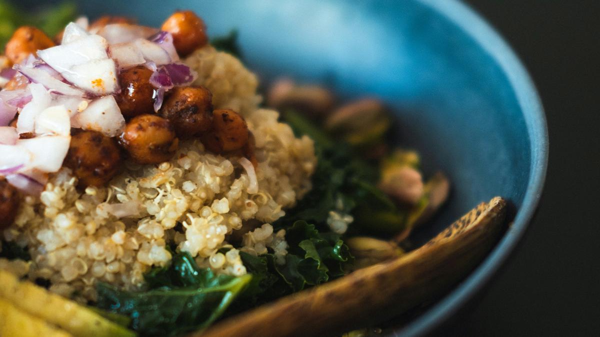 A blue bowl with quinoa, chickpeas, chopped onions, greens and a wooden spoon on a dark background.