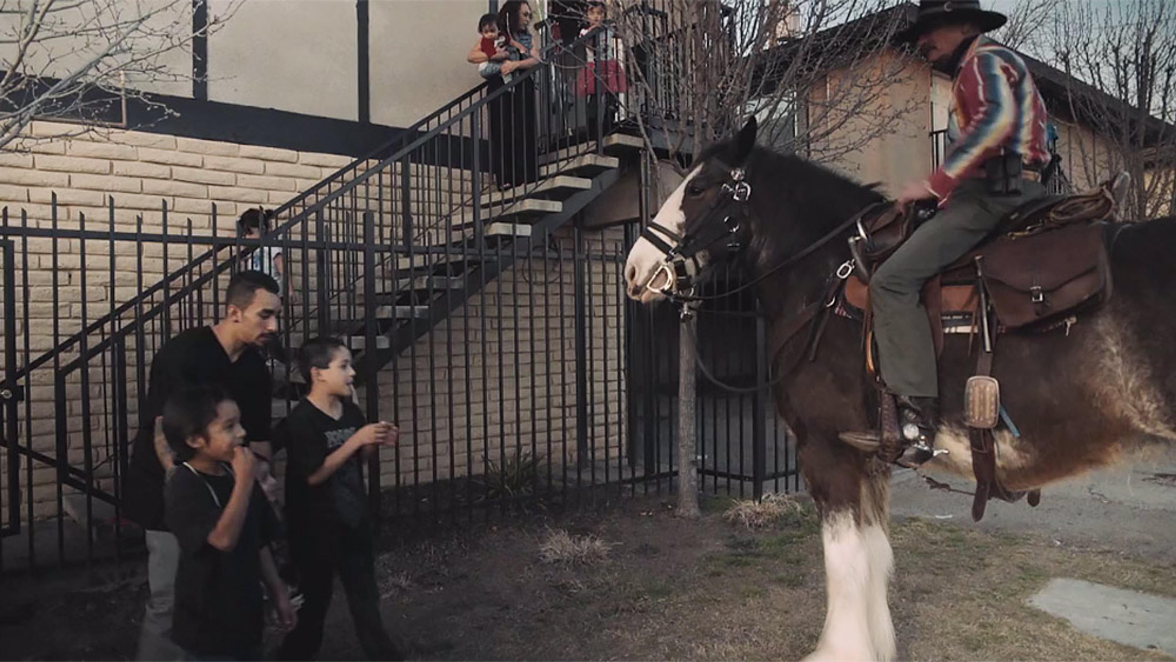 A man on a horse talking to children near a staircase. The scene is outdoors by a fence and a building in the background.
