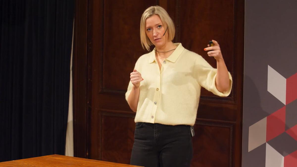 A woman with a headset giving a presentation. She gestures with her hand. The background is wood paneling.