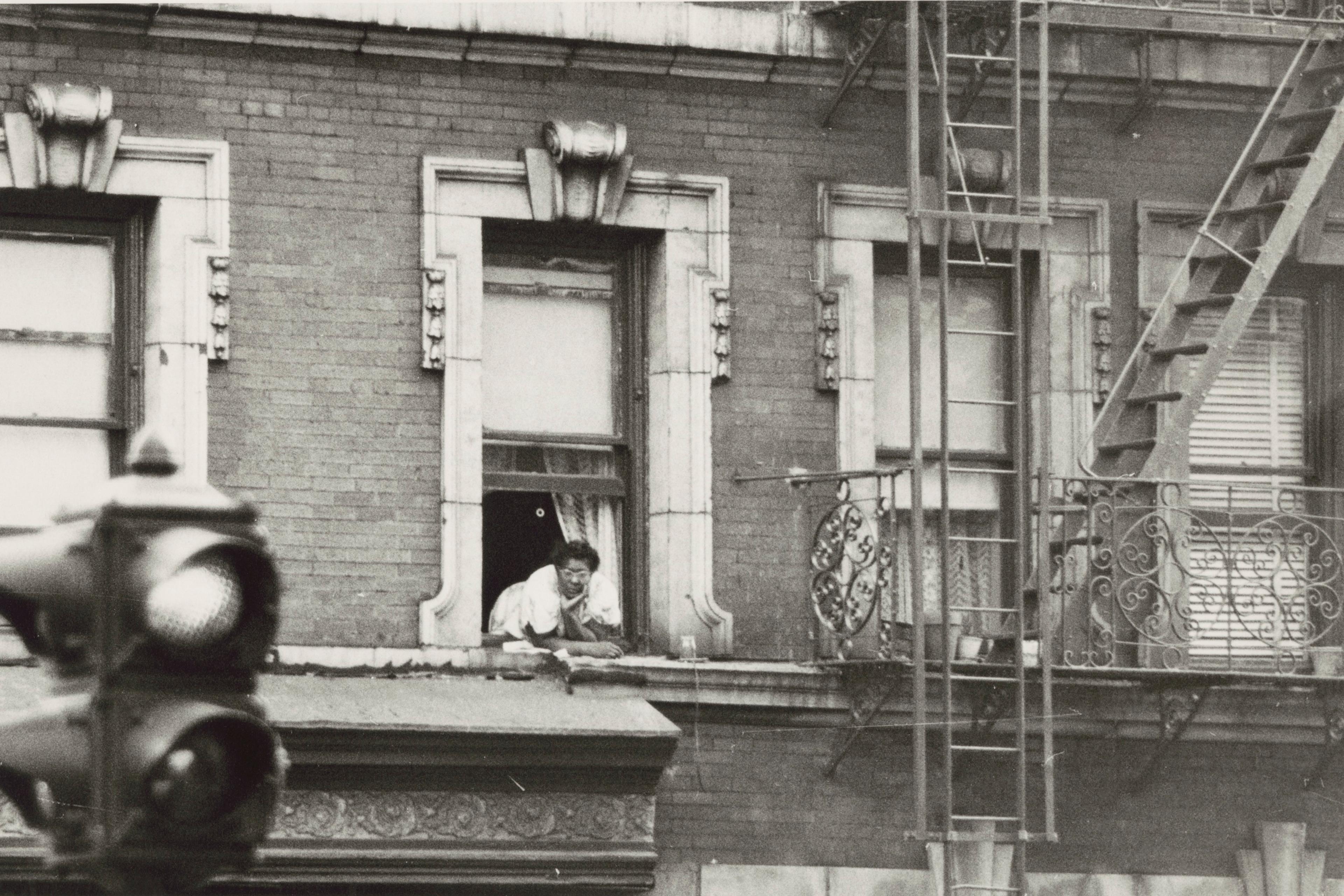 Black and white photo of a person leaning from a window in a brick building with a fire escape, traffic light in foreground.