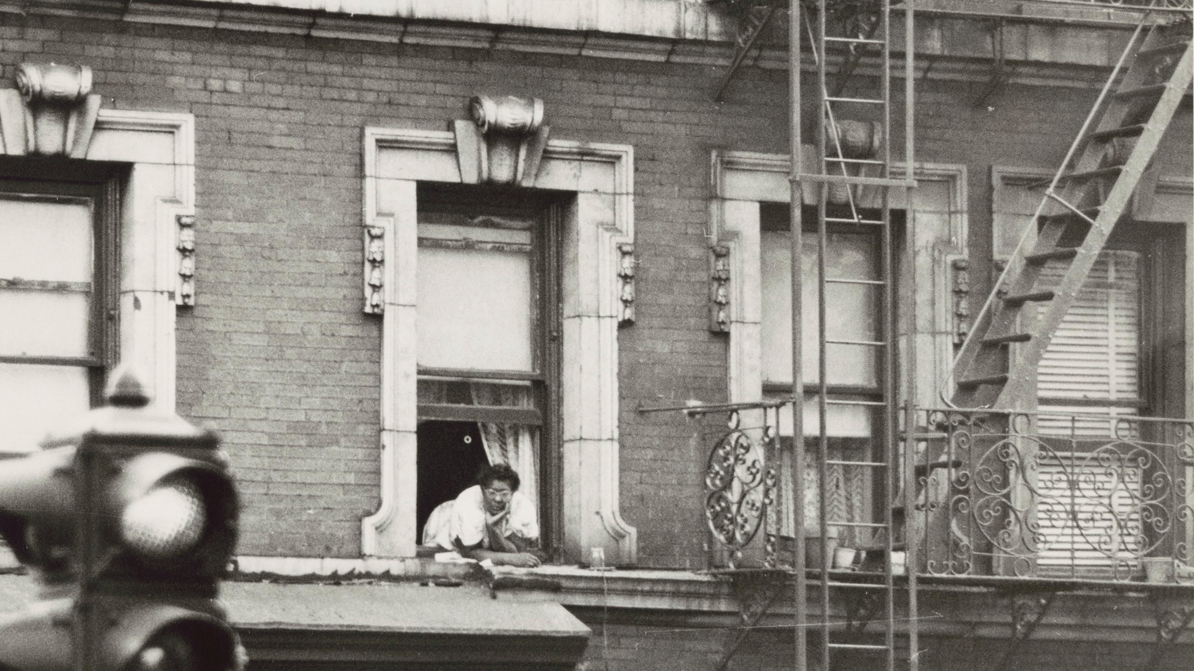 Black and white photo of a person leaning from a window in a brick building with a fire escape, traffic light in foreground.